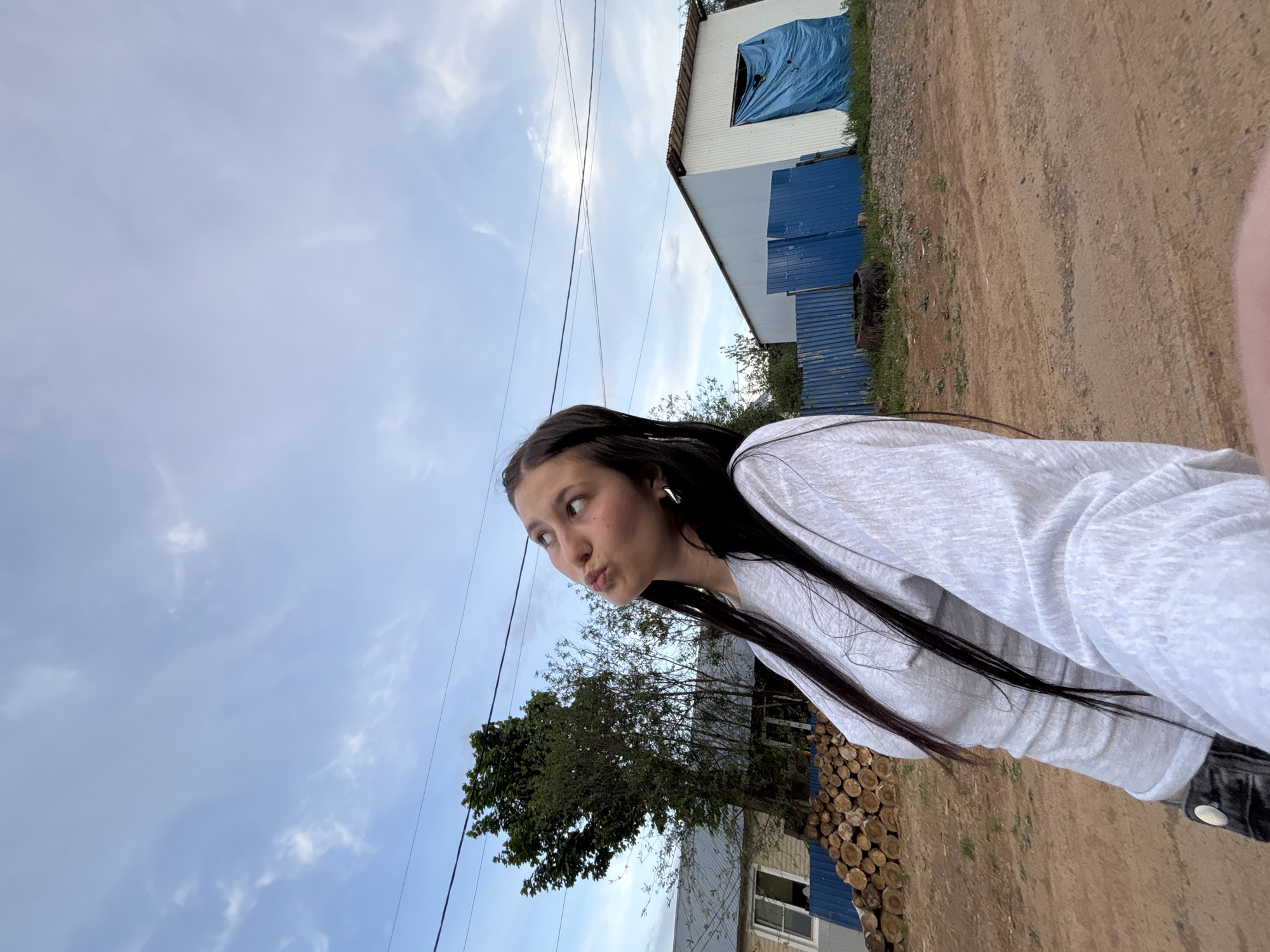 A young woman stands outside a building in Фоки, Russia. She has long, dark hair and is wearing a light gray shirt. She is looking to the left with pursed lips. Behind her is a small tree and a stack of logs, a partially visible house with a blue trim, a dirt road, and another building with blue tarp covering a window. The sky is cloudy. The photograph is tilted.