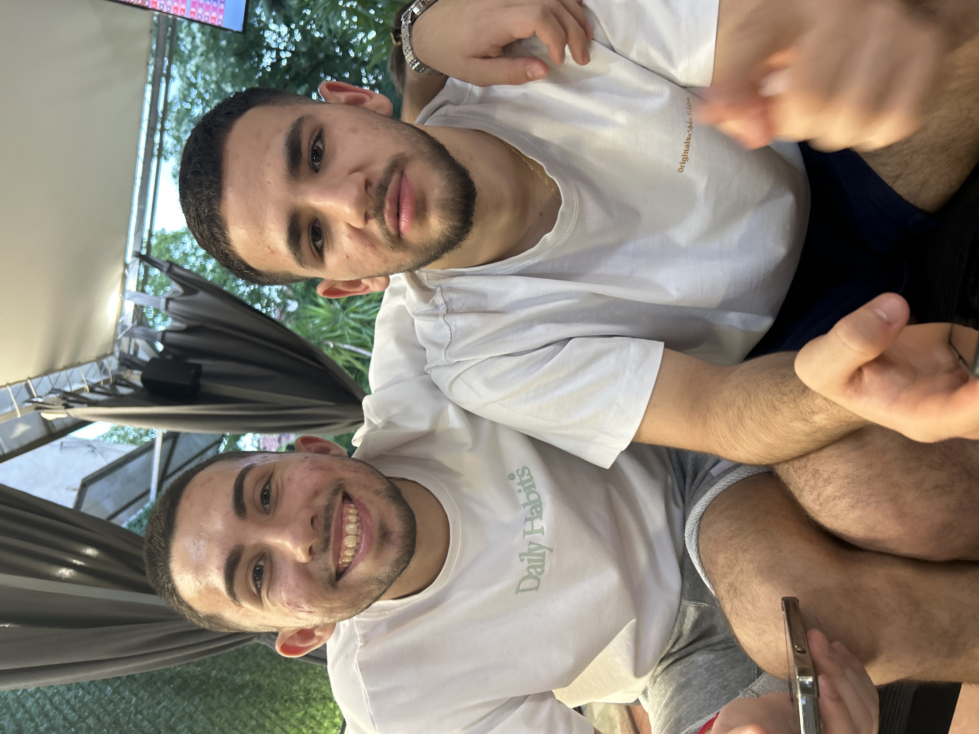 A full-length frontal shot captures two young adult males seated together in an outdoor or semi-outdoor setting in Blagoevgrad, Bulgaria, likely a cafe or restaurant patio. The man on the left, with dark, short hair and a trimmed beard, smiles broadly, revealing his upper teeth. He wears a white crew-neck t-shirt with "Daily Habits" printed in light green on the chest and grey shorts. Visible skin blemishes are present on his face. His left hand is partially visible, holding a smartphone. The man on the right, also with dark, short hair and a trimmed beard, has a neutral expression. He wears a white crew-neck t-shirt with "Originals Studio Edition" printed in light brown on the chest, dark blue shorts, and a silver-toned watch on his left wrist. A thin gold chain is visible around his neck. His left arm is extended across the other man's right shoulder. His right hand is blurred, suggesting movement. Both individuals have visible body hair on their arms and legs. The background features dark grey fabric panels or curtains, some open to reveal lush green foliage and trees under bright daylight. A white awning structure is visible overhead, and a brightly lit, pixelated screen, possibly a display board, is faintly visible in the upper right quadrant of the image.