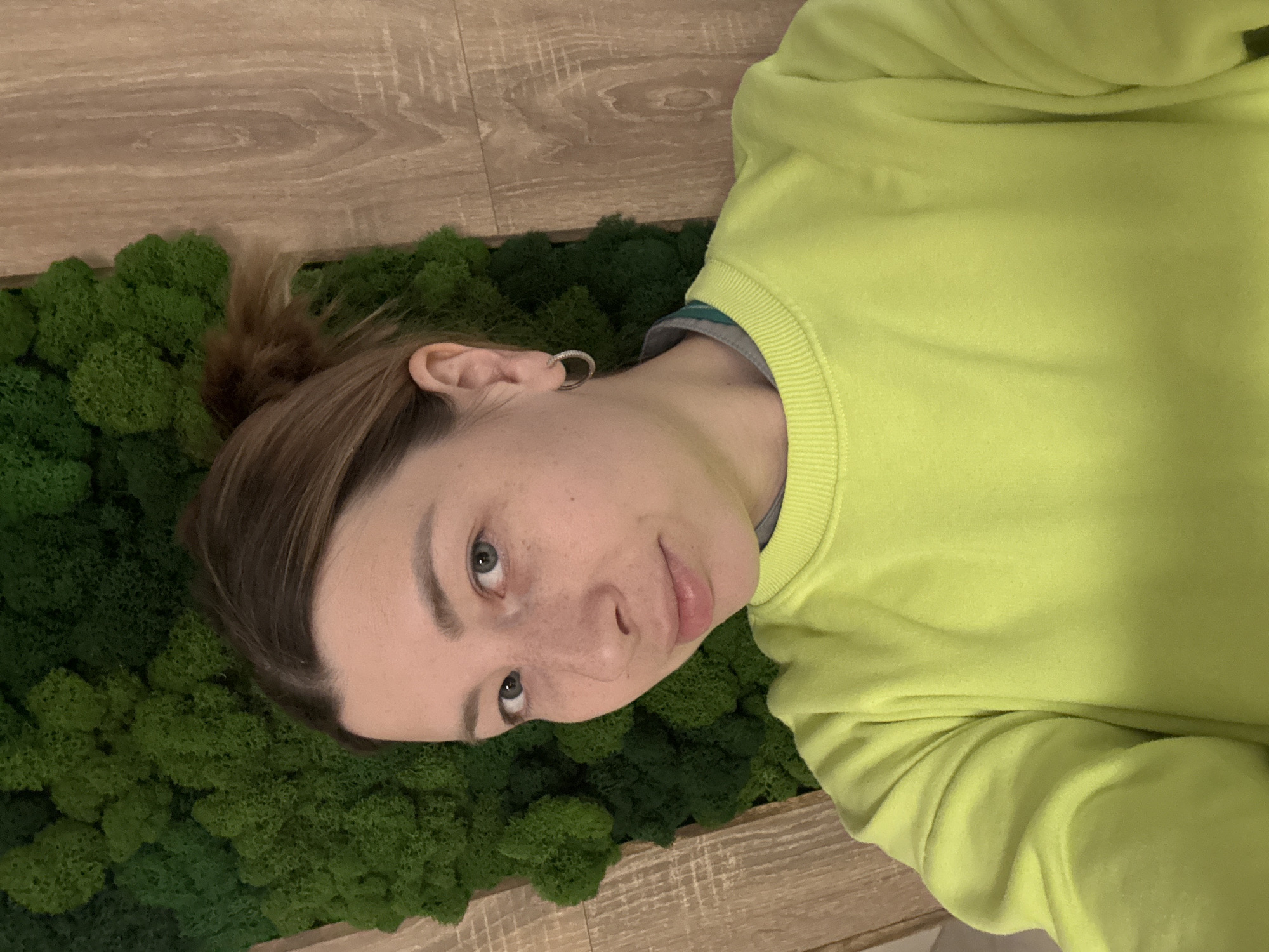 A person is lying down on a patch of green moss, which is situated on a wooden surface. The person is wearing a bright yellow-green sweater and has their hair pulled back in a bun. They are looking directly at the camera. No information on the location is given, and Moscow, Russia, is not discernible.