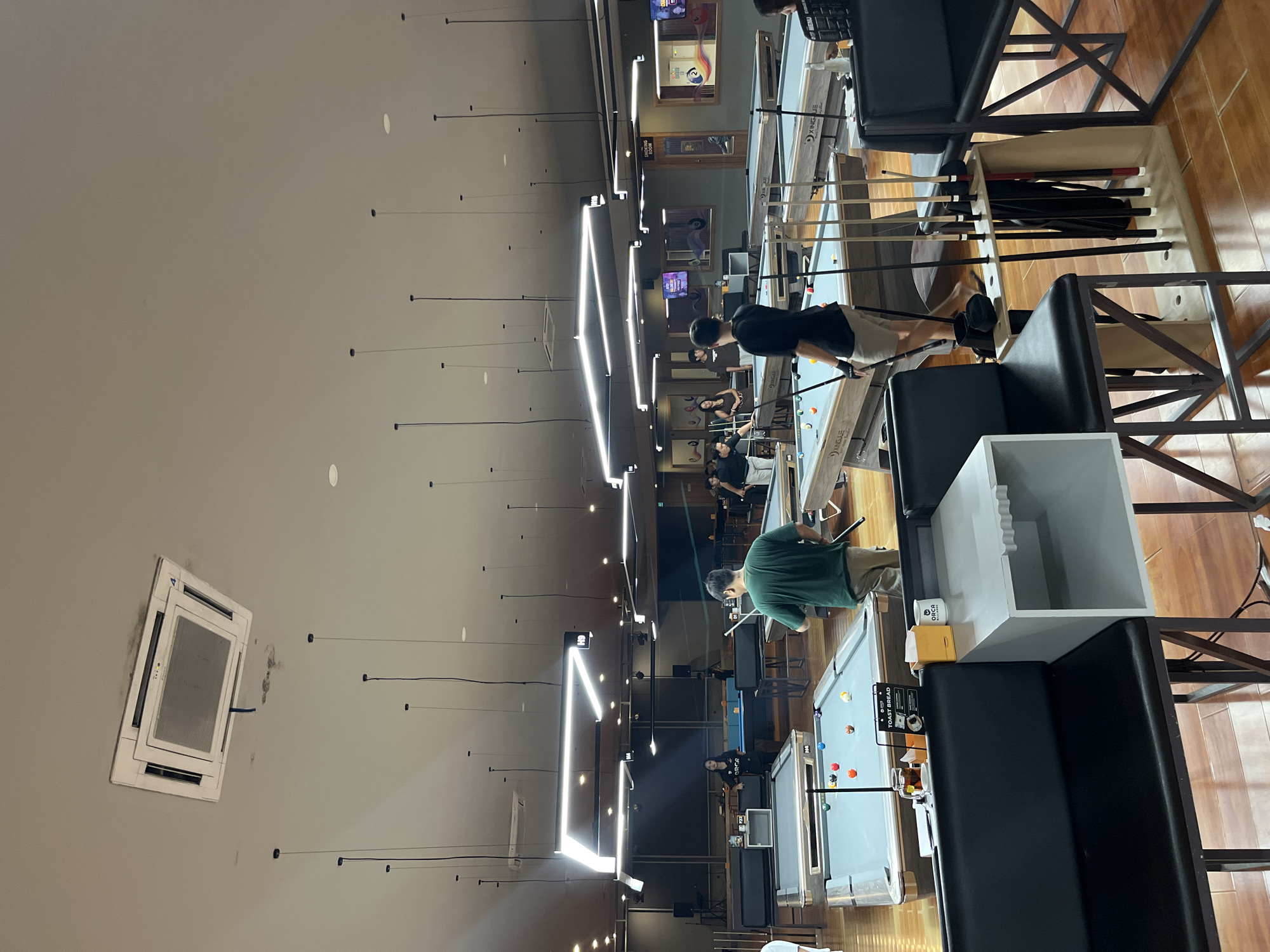 This media file shows a group of people playing billiards in what appears to be an indoor pool hall. The scene is brightly lit by modern, geometric LED light fixtures on the ceiling, suggesting it might be evening or indoors during the day. Several pool tables are visible, set up for games. People of various ages are engaged in playing, observing, or preparing to play. Some individuals are holding pool cues and are positioned at the tables, while others are seated on dark sofas lining the room, watching the games.
Notable objects include pool cues, billiard balls of various colors scattered on the tables, and a storage unit holding more pool cues. There's also a white cube-shaped object, possibly a table or storage, and an air conditioning unit mounted on a wall.
The environment has wooden flooring and a neutral-colored wall, creating a clean and contemporary aesthetic. There are framed pictures on the walls, adding a touch of decoration. The overall atmosphere seems relaxed and focused, as is typical for a billiards game.
The location is identified as Kuta Selatan, Indonesia. No specific time of day or weather conditions are discernible from the image itself, beyond the indoor lighting. No text is clearly legible enough to be transcribed.