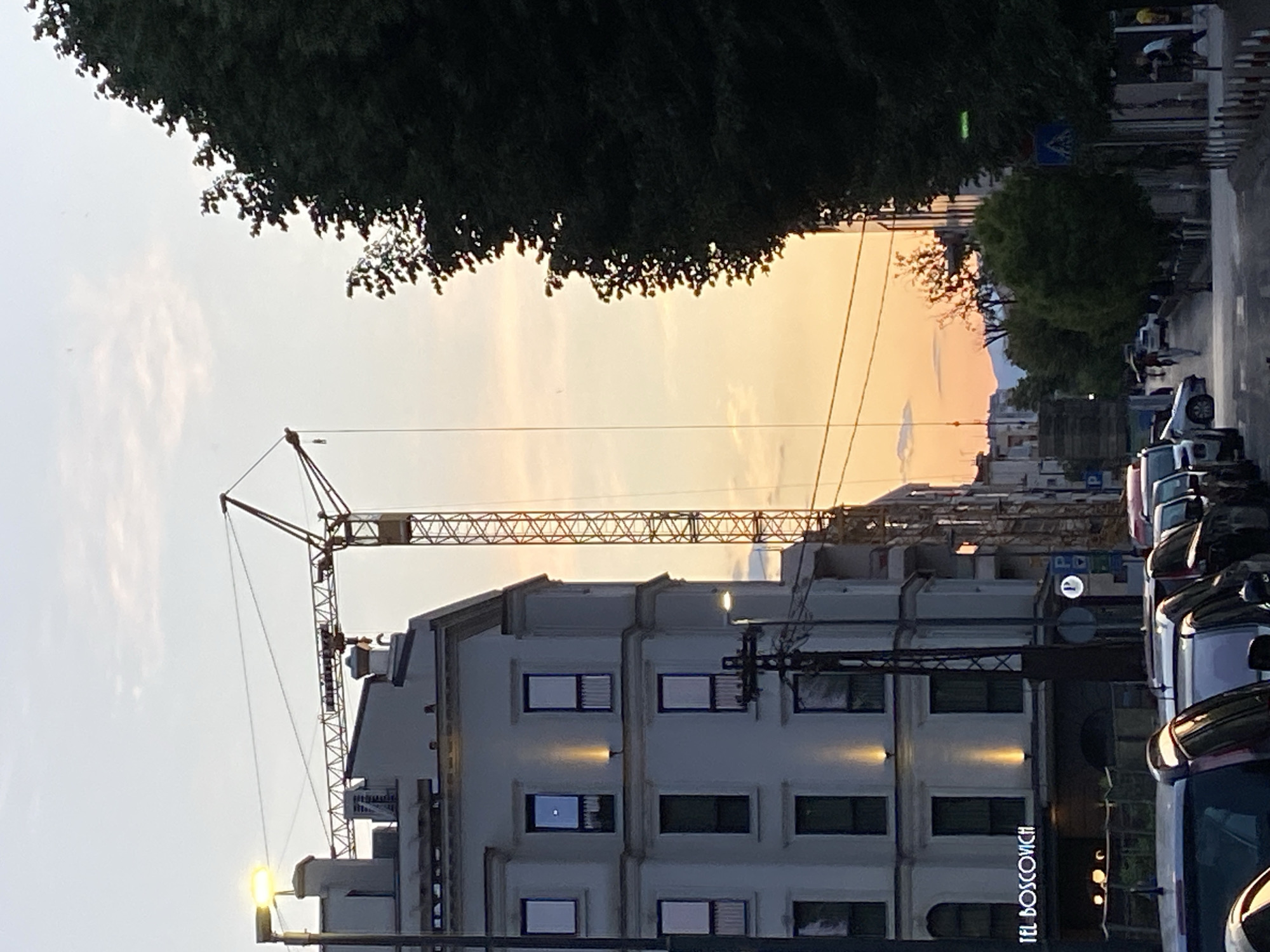 The image shows a street scene in Podgorica, Montenegro. A construction crane dominates the view above a multi-story white building with dark-framed windows. The sign “TEL BOSCOVICH” is visible on the building's facade. Cars are parked along the right side of the street. Trees line the street in the background, and the sky is a mix of light blue and orange hues, suggesting dawn or dusk. A pedestrian crossing sign is visible in the distance. There is a mountain range in the background.