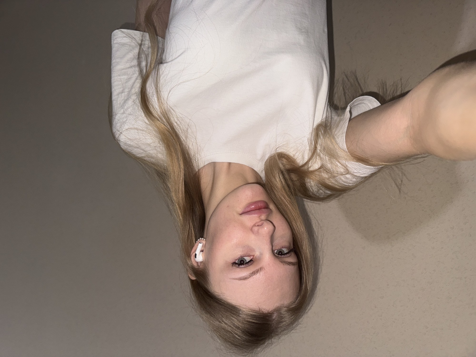 A blonde woman is pictured from a low angle. She is wearing a white t-shirt and a white earbud in her left ear. The background is a solid, light tan color. There is no indication of this image being in Perm, Russia.
