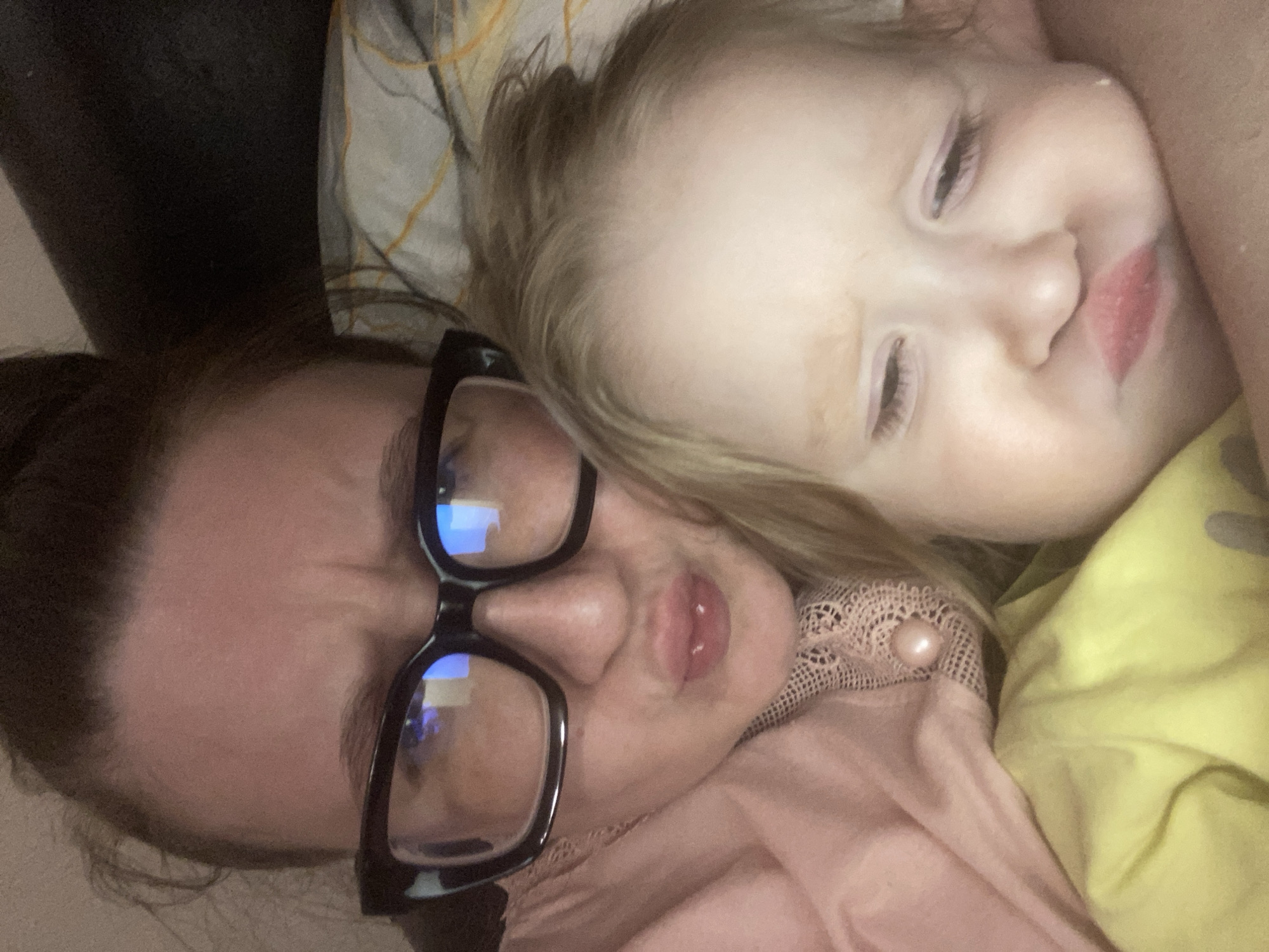 An indoor, close-up photograph taken in Chaikovsky, Russia, depicts two individuals: an adult female and a young child, positioned closely together. The adult female occupies the upper left portion of the frame, presenting dark hair pulled back, thick-rimmed black eyeglasses reflecting blue light, furrowed eyebrows, and pursed lips. She wears a light pink garment detailed with a lace trim and a single light-colored button resembling a pearl. The child, situated in the lower right, is nestled against the adult, exhibiting light-colored hair, partially closed eyes, and a similar pursed-lip expression. A faint streak is visible on the child's right cheek. The child wears a bright yellow top. The immediate background shows a dark, solid surface behind the adult and a patterned fabric, possibly a pillowcase or blanket, to the right, displaying light tones with abstract orange lines. The scene is illuminated by ambient, internal lighting.