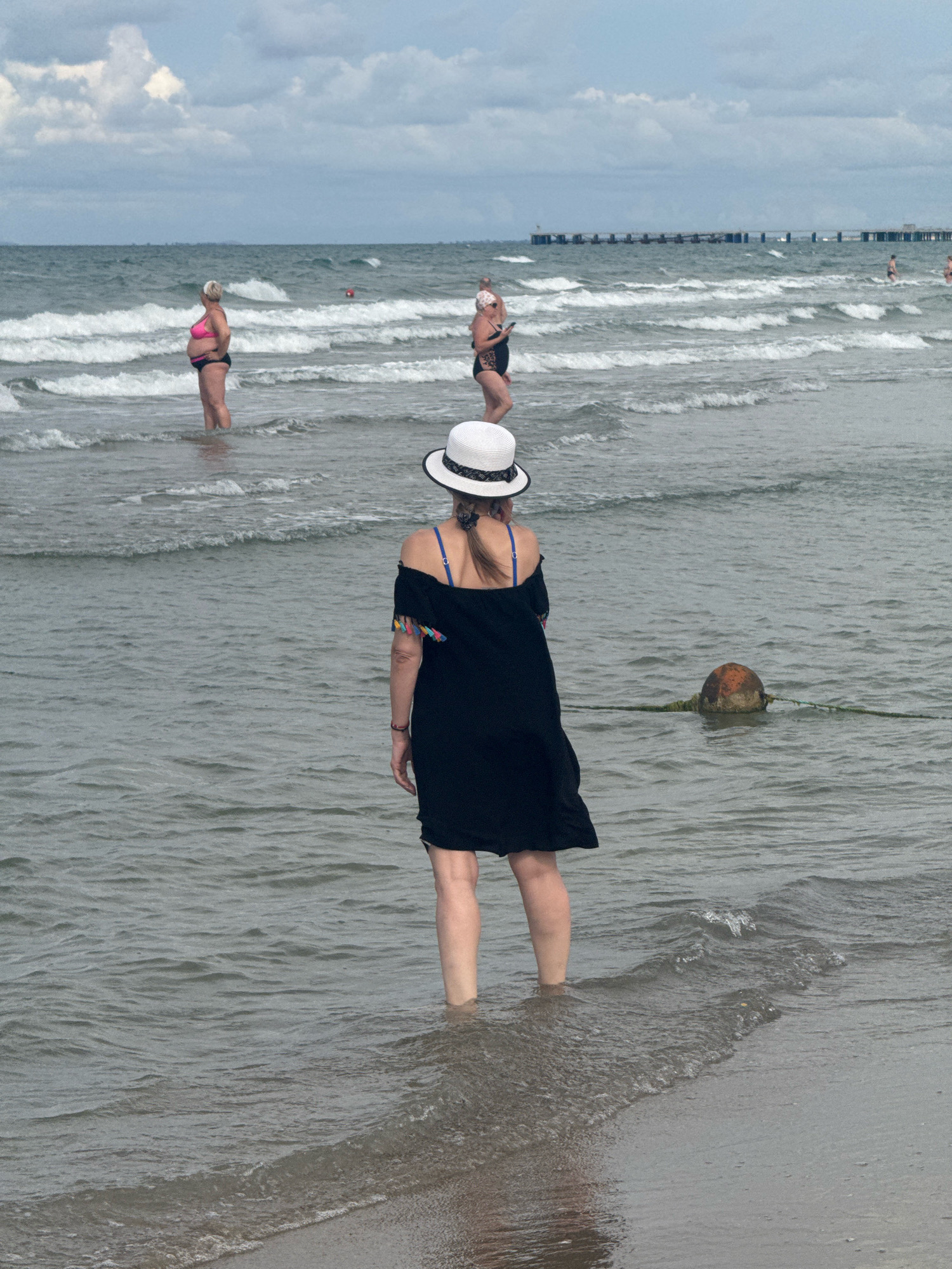 A woman wearing a black off-the-shoulder cover-up over a blue bikini top and a white hat stands in the shallow water of a beach, facing away from the camera. Her legs are submerged up to her calves. In the background, other people are visible in the water, and a pier extends into the sea. The water is choppy with small waves. A buoy is visible in the water to the right of the woman. The sky is overcast with clouds. The scene is likely the Black Sea coast in Anapa, Russia.