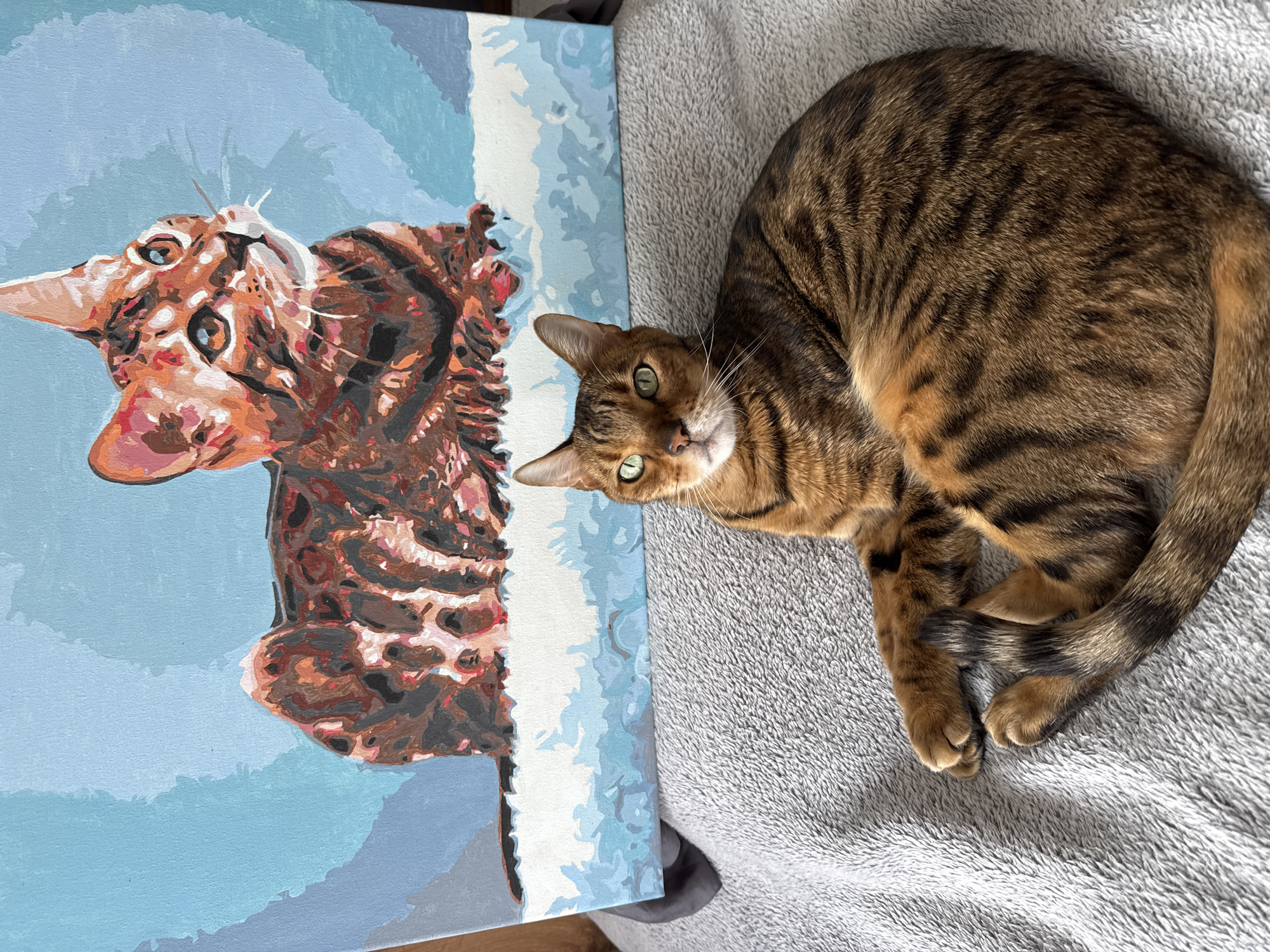A domestic cat with striking green eyes and a brown tabby coat with black stripes is prominently featured on the right side of the image. The cat is curled up on a soft, light grey textured blanket, looking directly towards the viewer with an attentive expression. Its front paws are tucked in, and its tail is wrapped around its body.
To the left of the cat rests a painting on a canvas, depicting what appears to be the same cat. The painting is oriented vertically, with the cat's head towards the top. It exhibits a distinct paint-by-numbers style, characterized by segmented areas of color including various shades of brown, orange, black, and white for the fur, and bright green for the eyes. The painted cat's form is set against a light blue background with some cloud-like white and darker blue elements. The painting is propped up, possibly leaning against a surface.
The scene is brightly lit, suggesting it is daytime. The setting appears to be indoors, with the grey blanket suggesting a comfortable, domestic environment. There is no visible text in the image.