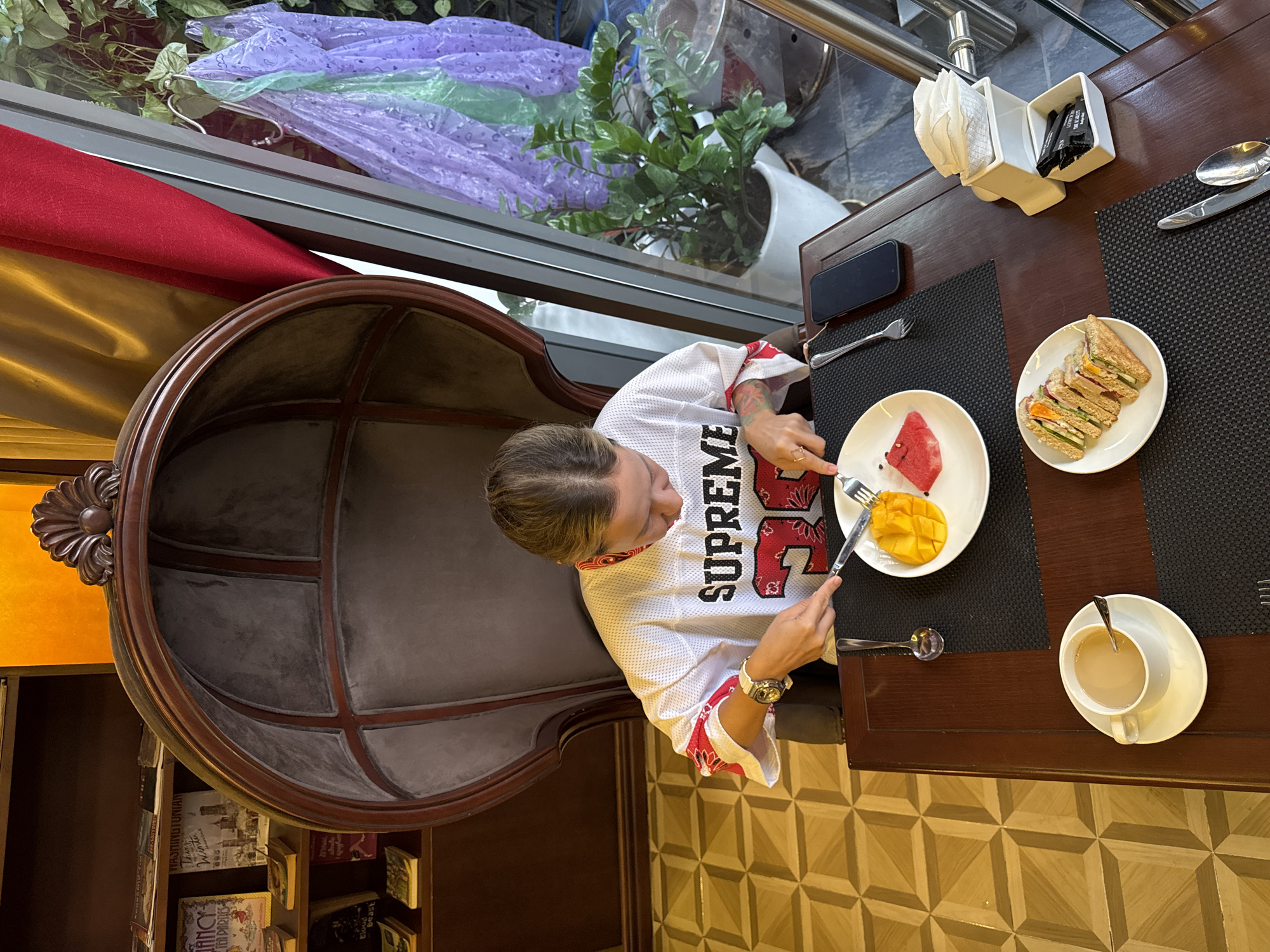 In Thuận Hóa Ward, Vietnam, a young woman with blonde hair pulled back is seated in a large, dark brown, egg-shaped chair with upholstered gray seating. She wears a white jersey-style shirt with red and white patterned sleeves and the number "36" on the front. The woman is using a fork and knife to eat fruit from a white plate containing a slice of watermelon and several pieces of mango. A cup of coffee sits on a saucer to her left, and a plate of sliced sandwiches is positioned further to the left on the table. A black smartphone is visible on the table to her right. The scene appears to be indoors, with bookshelves visible in the background and a large window to the right showing greenery outside and purple and green plastic coverings hanging. The floor is tiled with a geometric pattern.