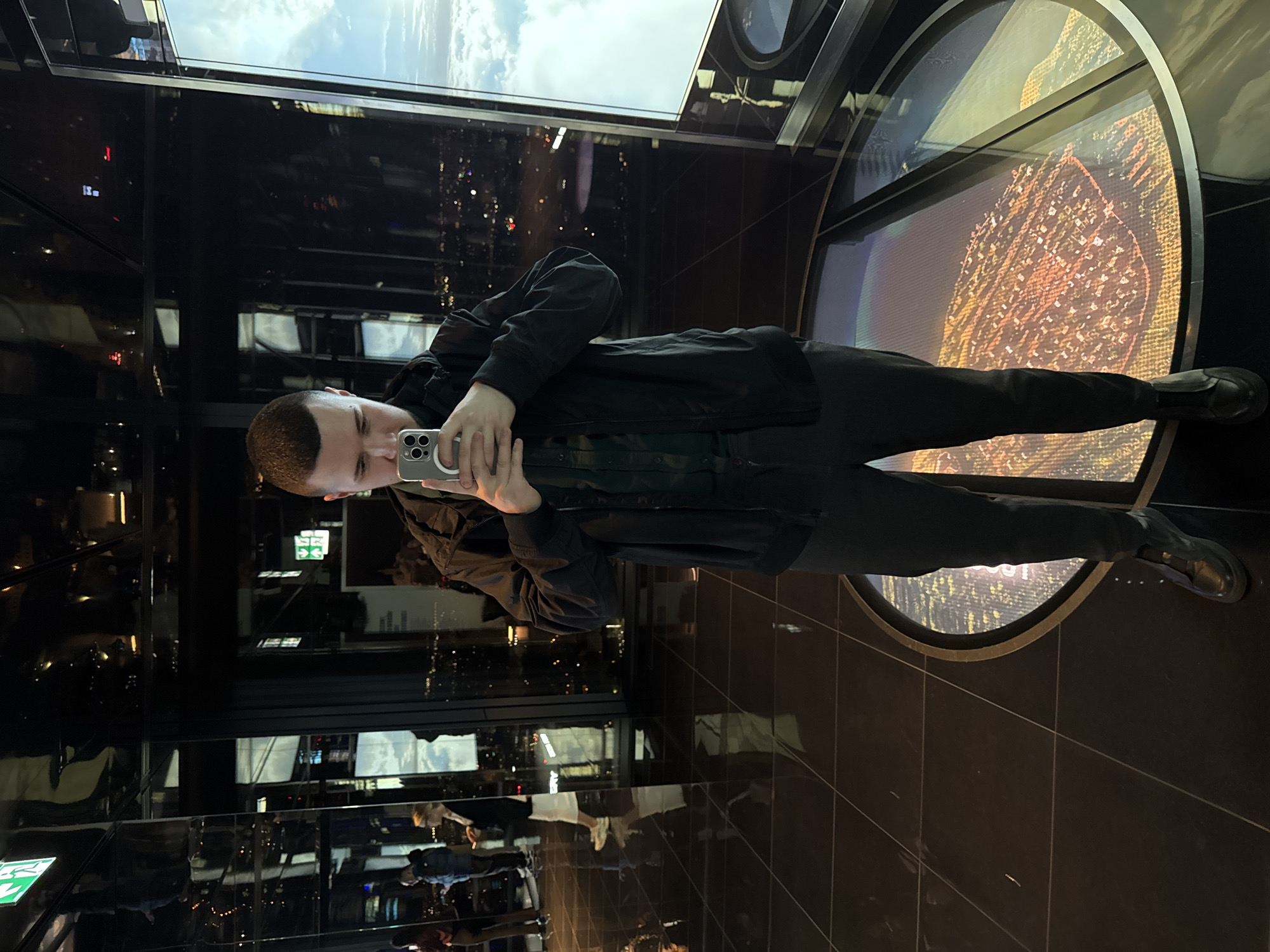 A person is standing in an observation deck in Warsaw, Poland, holding a smartphone and taking a picture. The deck is on a high floor, and it has a large, circular window in the floor. The city lights and skyline are visible through the window. The person is wearing dark clothing and is standing with their feet apart.