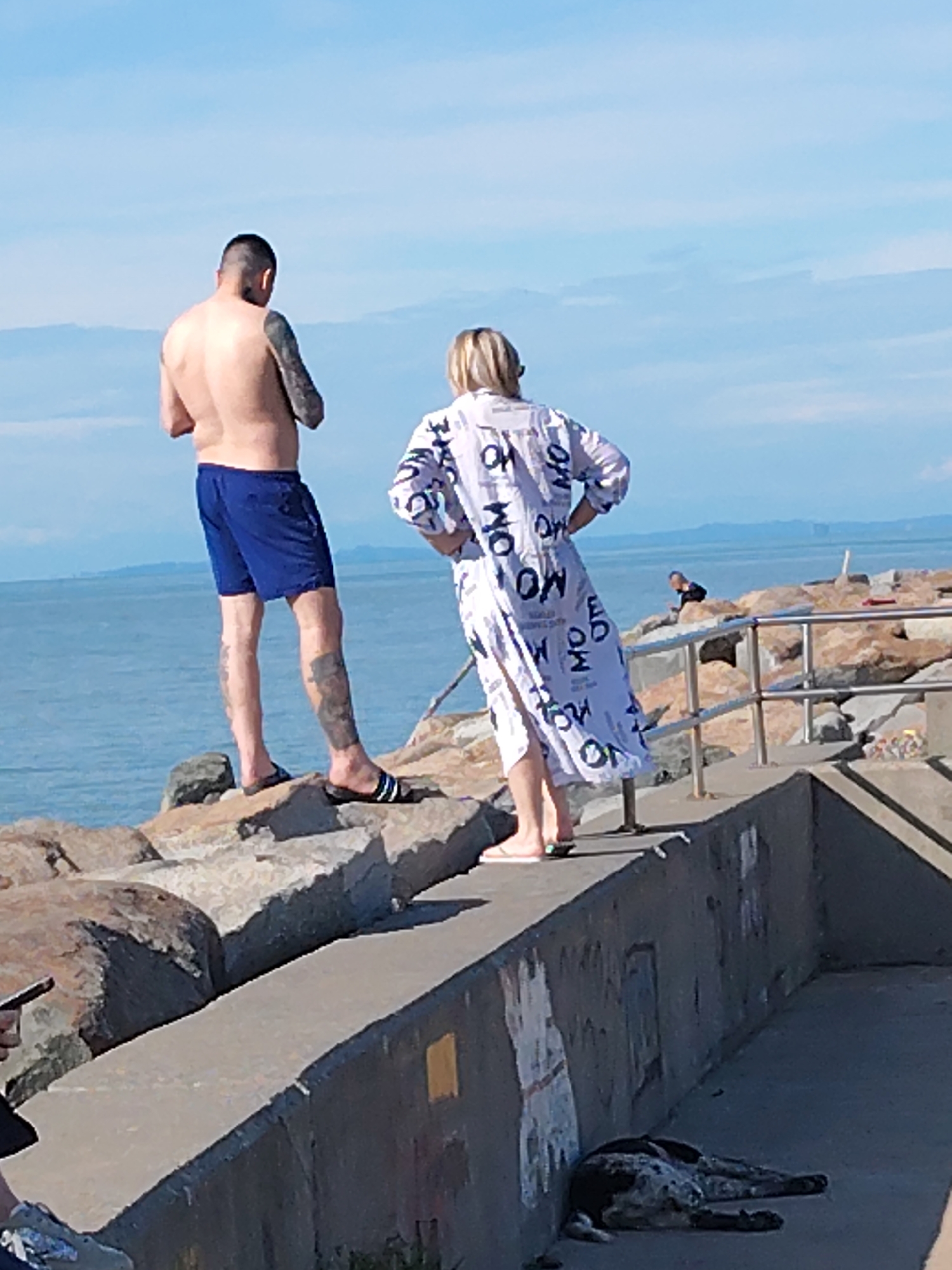 Two people, a man and a woman, are standing on large rocks along the shore in Batumi, Georgia. The man is shirtless and wearing blue swim trunks and black and white sandals, and he has tattoos on both arms and one leg. The woman is wearing a white, knee-length robe with a pattern of black text. A dark-colored dog is lying on the ground beneath a concrete wall. In the background, the Black Sea stretches out to the horizon under a blue sky. A third person is sitting on the rocks beyond the woman. A concrete barrier with a metal railing is between the woman and the person.