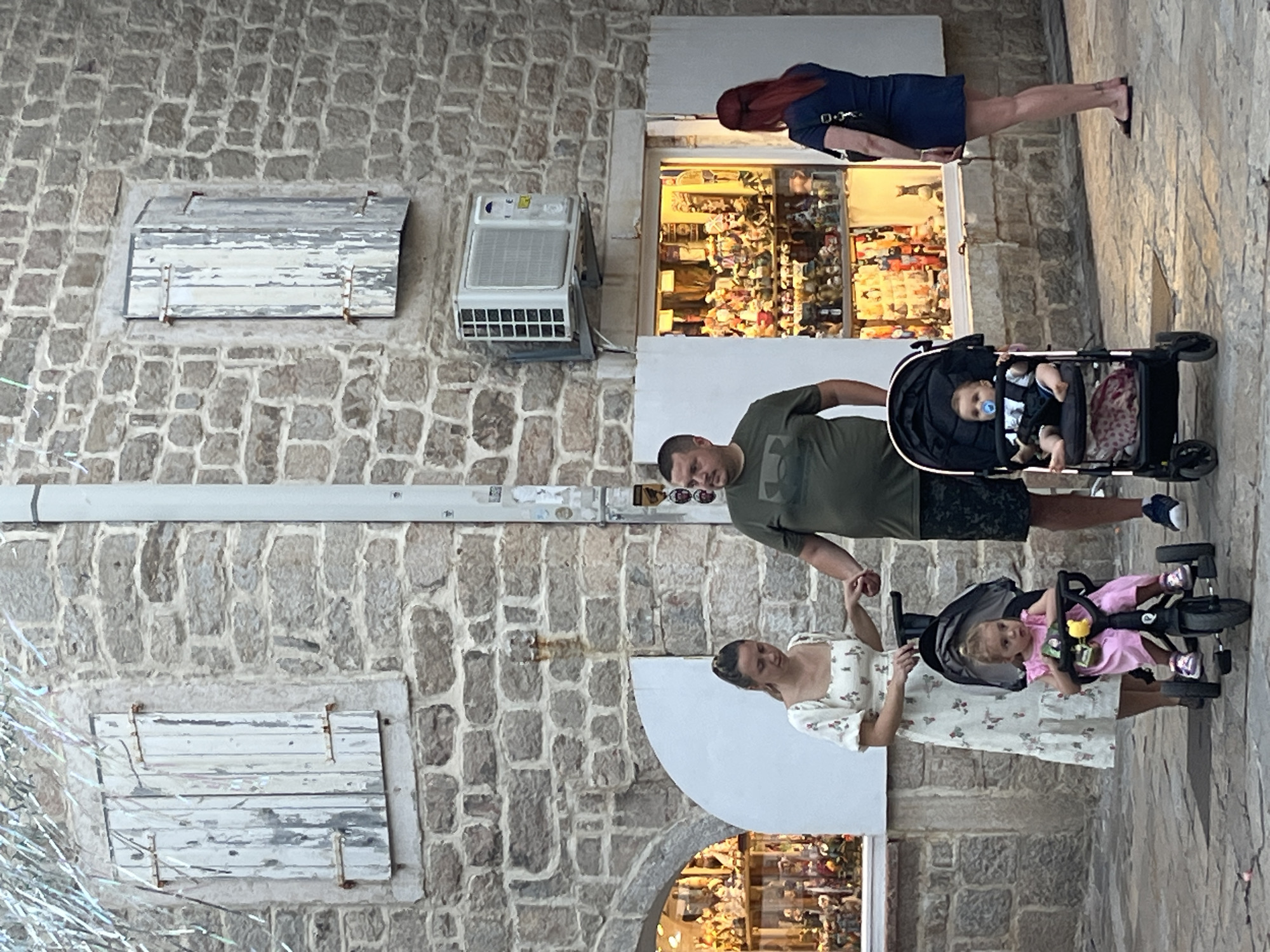 This is a medium-shot, eye-level outdoor photograph of a family with two young children in strollers. A couple, a man and a woman, are standing in the foreground, holding hands. The woman is wearing a light-colored, floral-print dress. The man is wearing a dark green t-shirt and patterned shorts. They are standing next to two strollers. The stroller on the left contains a young girl wearing a pink dress and white sneakers. The stroller on the right contains a baby with a pacifier. The background features a stone building with two shuttered windows. An air conditioning unit is mounted on the wall above a shop window filled with merchandise. A woman with red hair wearing a dark blue dress stands to the right of the shop. The scene appears to be on a cobblestone street in Budva, Montenegro, possibly near the Old Town.