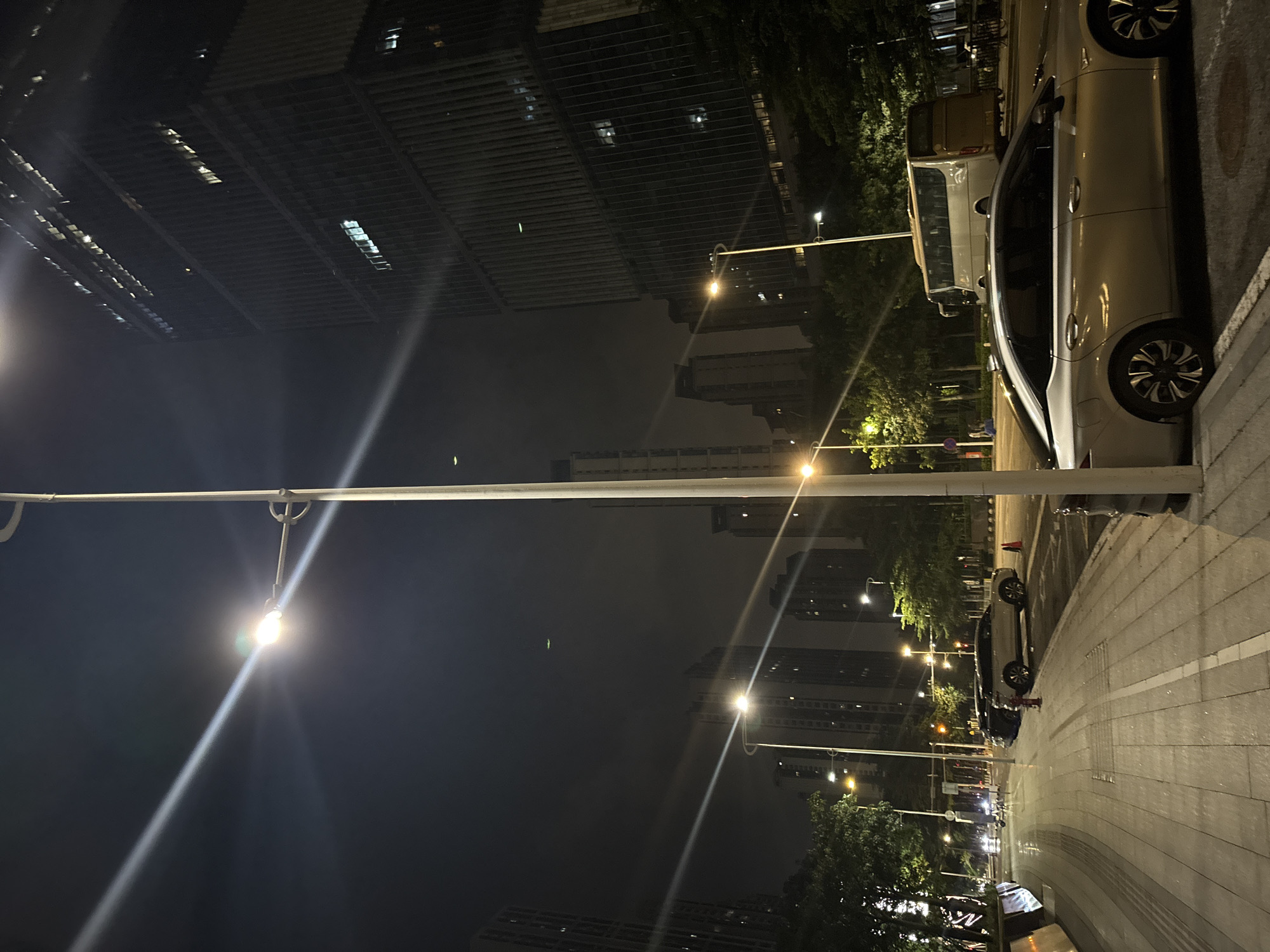 This nighttime image captures a street scene in Haizhu District, China. The perspective is low, looking upwards at tall buildings and streetlights. A light-colored car is parked on the right side of the frame, its sleek design and alloy wheels visible. Behind the car, a light-colored van is also parked. The street appears to be paved with interlocking bricks, and there are trees lining the sidewalk, their leaves illuminated by the artificial lights.
Several streetlights are visible, some with a warm yellow glow and others casting a bright white light. One streetlight, in the foreground, is particularly prominent, with light rays radiating outwards. The buildings in the background are modern, with many windows reflecting the city lights. The sky is dark, indicating it is nighttime. The atmosphere feels urban and contemporary. There is no visible text in the image.