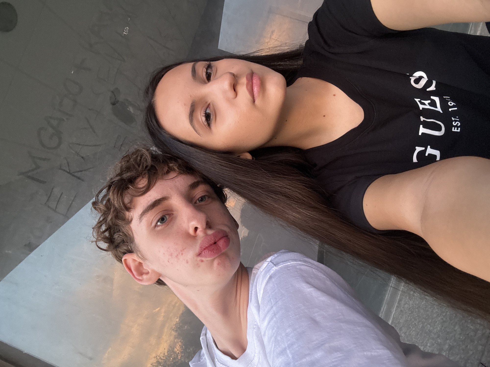 Two individuals, a male and a female, are captured in a selfie. The male, on the left, has light skin, curly brown hair, and blue eyes. He is wearing a white t-shirt and has his lips pursed. The female, on the right, has dark hair and brown eyes, and is wearing a black t-shirt with the word "GUESS" printed on it. They are positioned in front of a reflective surface, possibly a window or a glass building, which shows a soft, golden light, suggesting it might be sunset or sunrise. Some graffiti is visible on the surface behind them. The background also shows some grey, textured material, likely a wall or pavement. The overall scene appears to be outdoors, in an urban setting, given the graffiti and building elements.