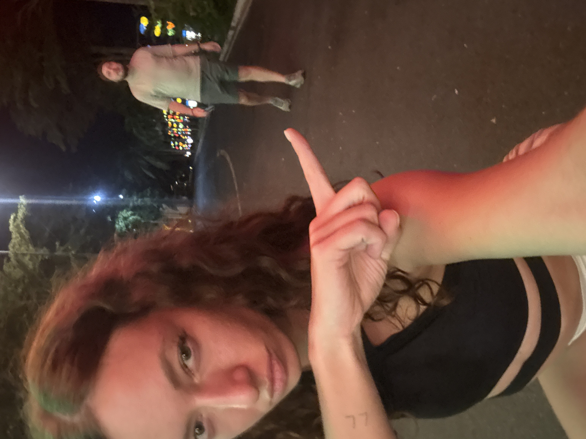 A low-angle, nighttime photograph depicts a woman pointing her index finger towards the upper left corner. She has fair skin, dark curly hair, and is wearing a black crop top with mesh panels. A small tattoo is visible on her inner forearm. In the background, a man is standing on a street, wearing a light-colored shirt and dark shorts. He appears to be walking away from the camera. Strings of colorful lights are hanging from trees in the background, indicating a festive atmosphere or event. The scene is located in Hội An Ward, Vietnam, based on the string lights being located in Hoi An. The ground is paved, and the lighting is dim overall.