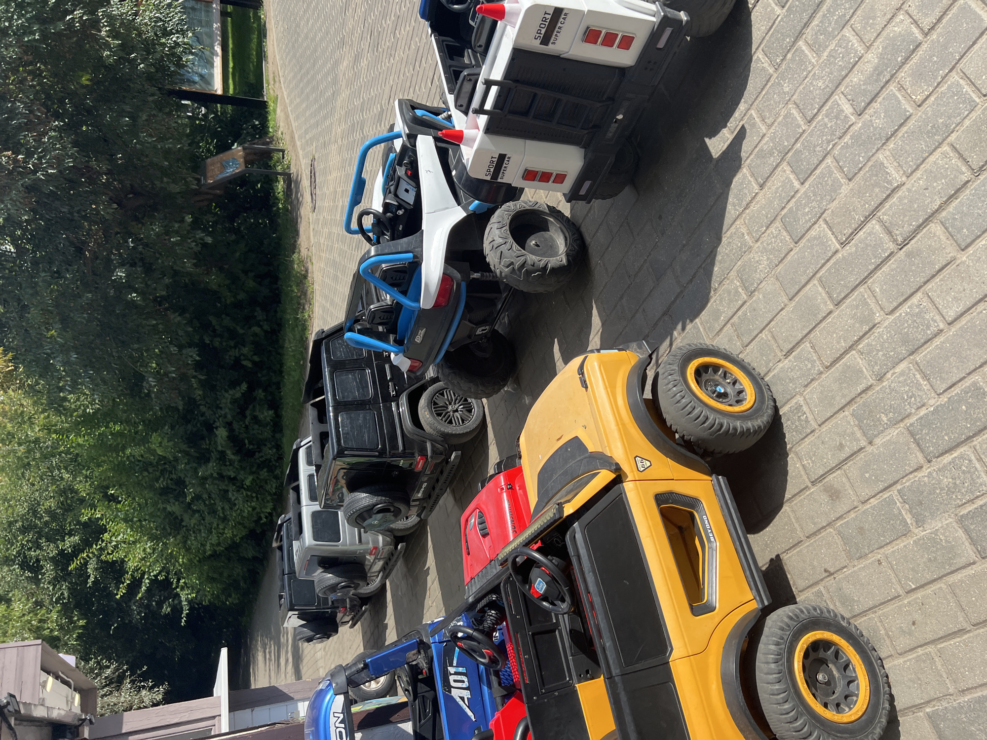 The image shows a row of toy electric vehicles, likely for rent or sale, parked on a paved area outdoors. The vehicles include several black and silver Mercedes-Benz G-Wagon style cars, a blue off-road vehicle with the number "401" on it, a red open-top vehicle, a yellow off-road vehicle, and two white and black UTV-style vehicles. The UTVs have "SPORT SUPER CAR" printed on them. The vehicles are parked in front of a building with a brown facade and a green, leafy background of trees and bushes. Sunlight casts shadows on the ground, indicating a sunny day. The paving stones are a light grey color. The scene appears to be a recreational area or an outdoor market. It is not possible to determine the specific location within Moscow, Russia, from the visual information provided. There are no people visible in the image.