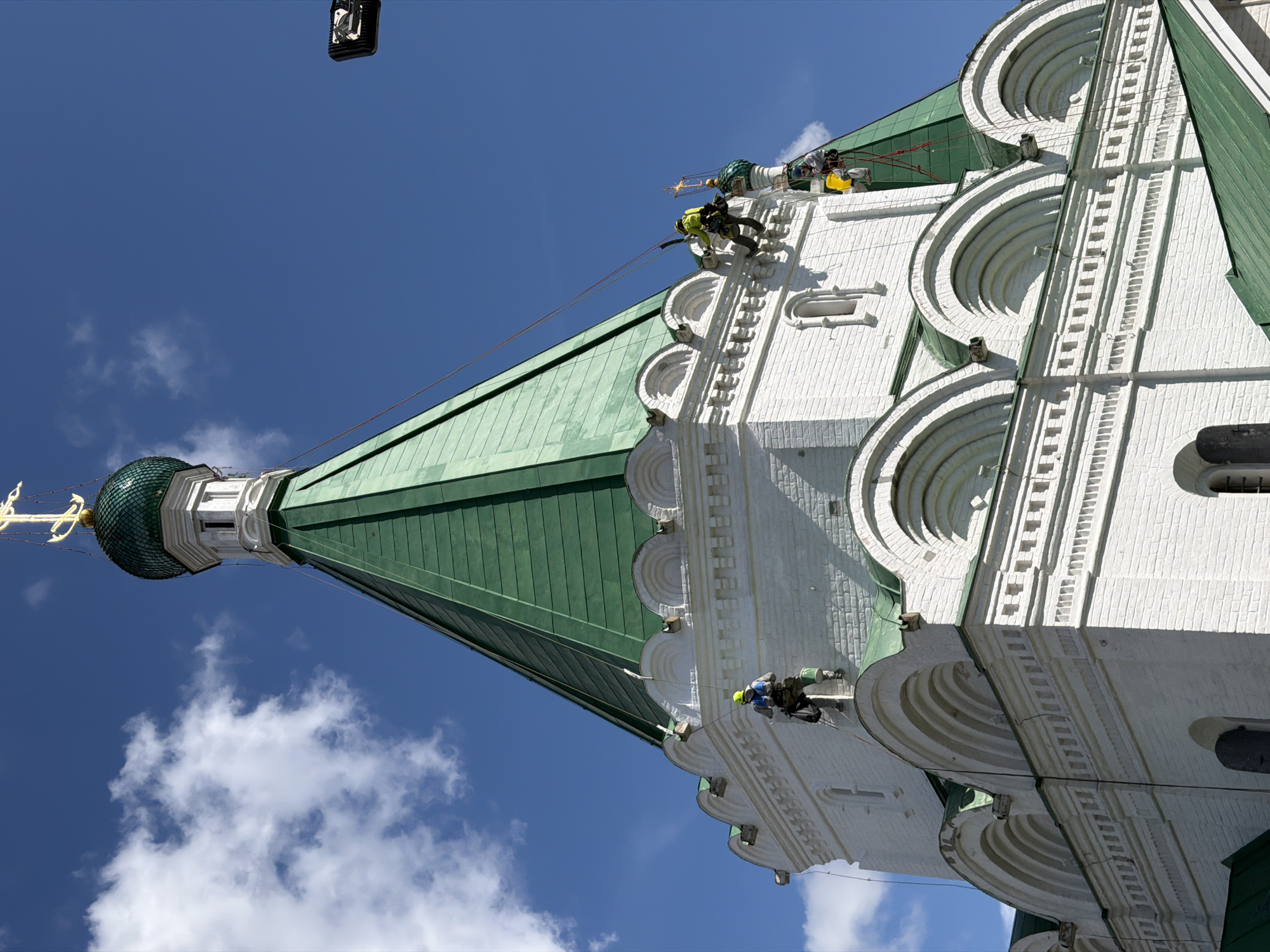 The image shows an exterior view of the Assumption Cathedral (also known as the Pechersky Ascension Monastery Cathedral) in Nizhny Novgorod, Russia. Several individuals, equipped with safety harnesses and ropes, are engaged in high-altitude work on the white brick facade and the green, tiered roof of the cathedral. One person is visible descending on a rope on the left side of the frame, another is perched on a ledge near the top, and a third is working higher up on the green roof. The bright blue sky with scattered white clouds serves as the backdrop, and a black object, possibly a camera or light fixture, is visible in the upper right corner against the sky. The architectural style of the cathedral is distinctly Russian, characterized by its onion domes and ornate detailing. The sun is shining, casting shadows and highlighting the textures of the building materials.