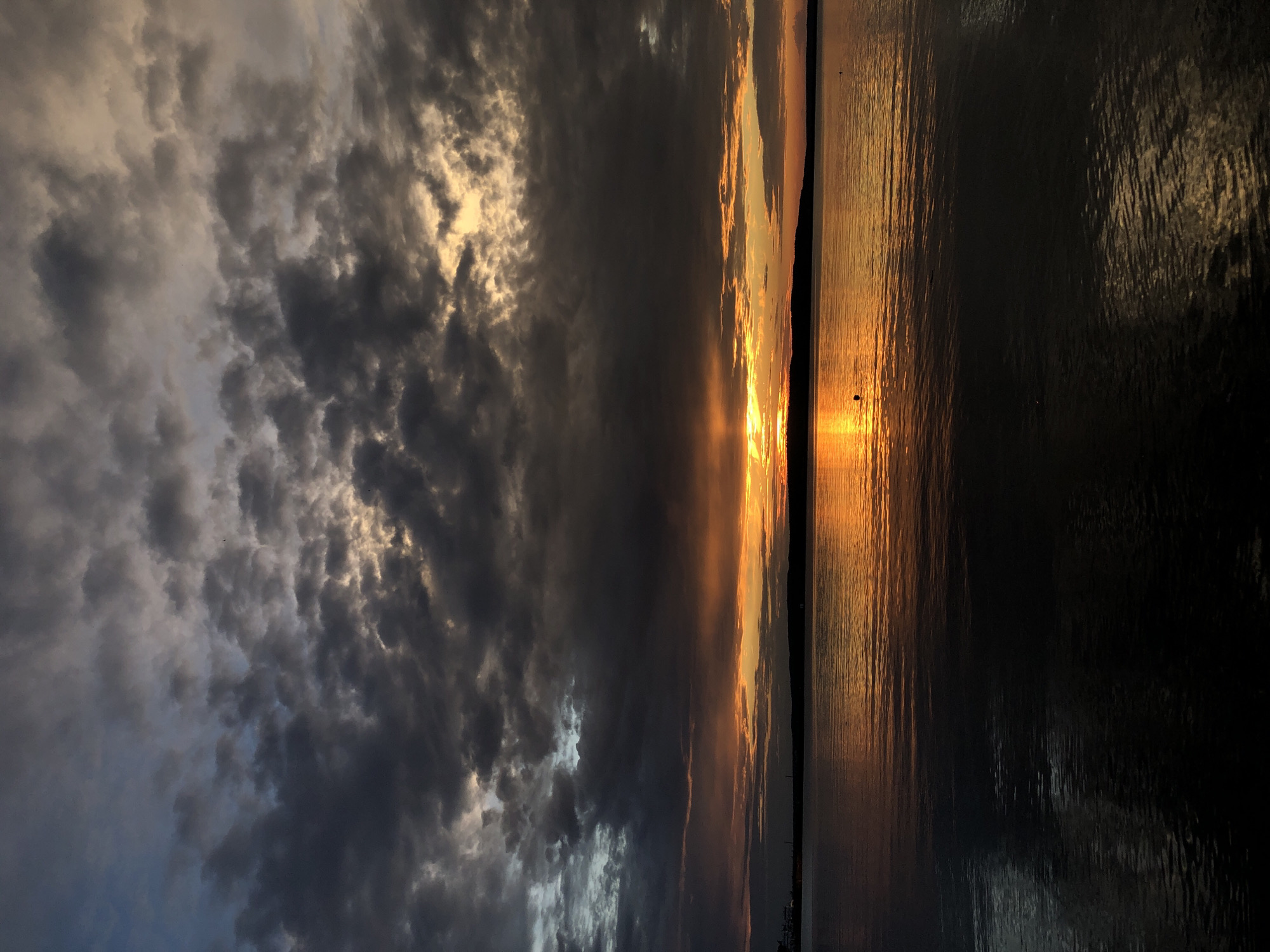 A dramatic sunset over the Kama River in Chaikovsky, Russia, is captured in this vertical, outdoor, full shot. The sky is a canvas of dark, brooding clouds interspersed with vibrant streaks of orange and yellow light, indicating the setting sun. The sun itself is a bright, concentrated orange ball peeking through a break in the clouds, casting a shimmering golden reflection on the calm water. The horizon is marked by a dark silhouette of a distant shore, possibly with some faint structures. The foreground is dominated by the dark, rippling surface of the river, reflecting the muted colors of the sky. There are two small, dark, spherical buoys visible in the water, one closer to the left and another further to the right. The overall mood is serene yet powerful, with the raw beauty of nature on full display.