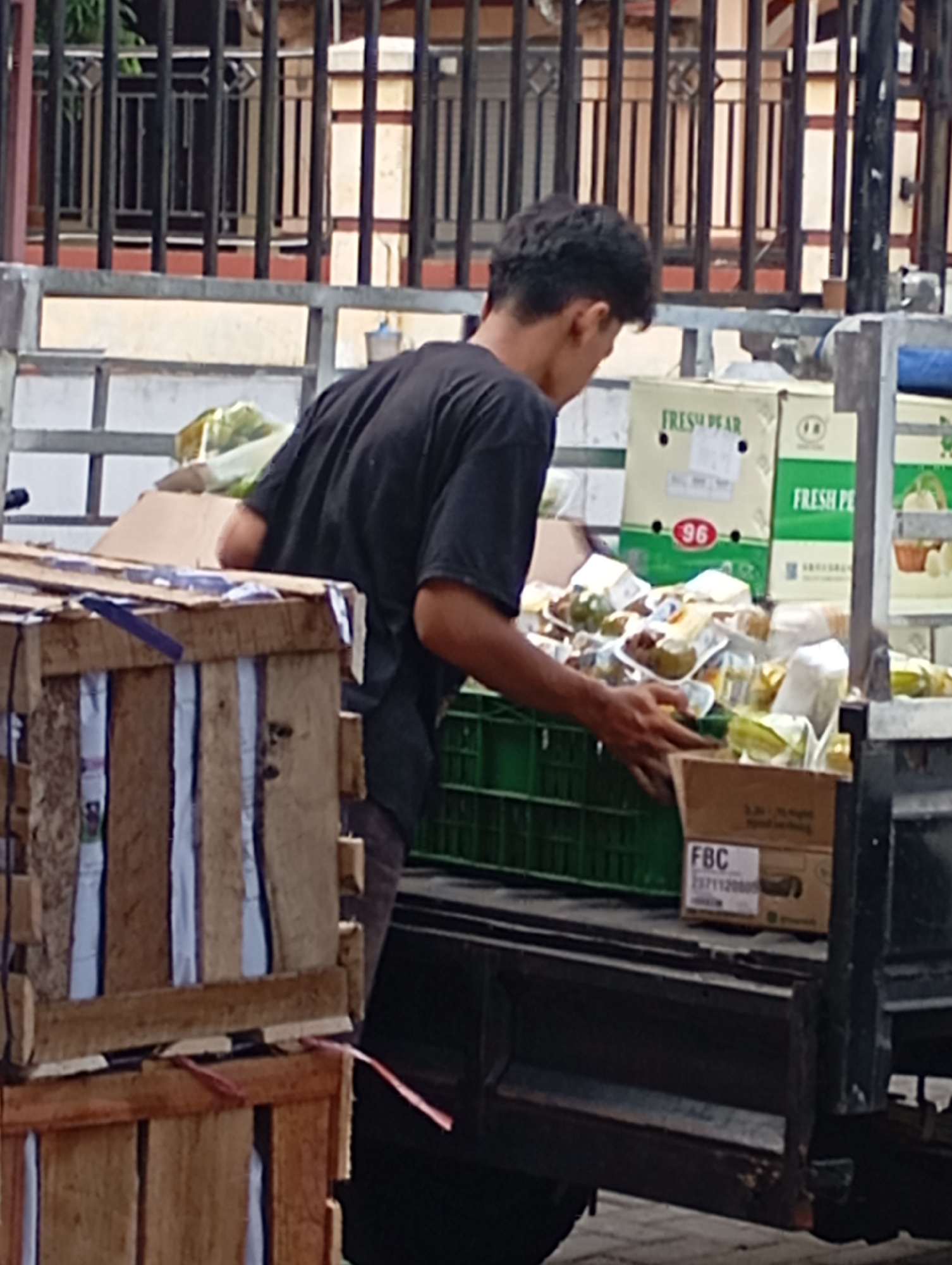A young male, wearing a black t-shirt, is seen from the back in the city of Plintahan, Indonesia. He is standing on the back of a truck laden with produce and wooden crates. The truck appears to be parked on a street, with a building and fence in the background. The man is in the process of unloading or organizing the goods, as his hands are near a green plastic crate filled with packaged fruits and vegetables. Several cardboard boxes are stacked on the truck, some with labels like "FRESH PEAR" and "FBC". The foreground is dominated by wooden crates, one of which is partially visible. The overall scene depicts a delivery or distribution of produce.