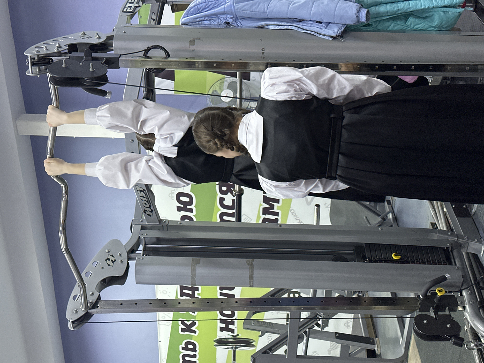 Two young women, possibly students, are using a weight-training machine. They are both wearing white blouses and black skirts/dresses, suggesting a school uniform. The machine is located in a gymnasium in Votkinsk, Russia, as indicated by the Russian text on the machine. The machine is silver, and there are visible weights and pulleys. The background is a light blue wall. There are also some folded clothes on top of the machine.