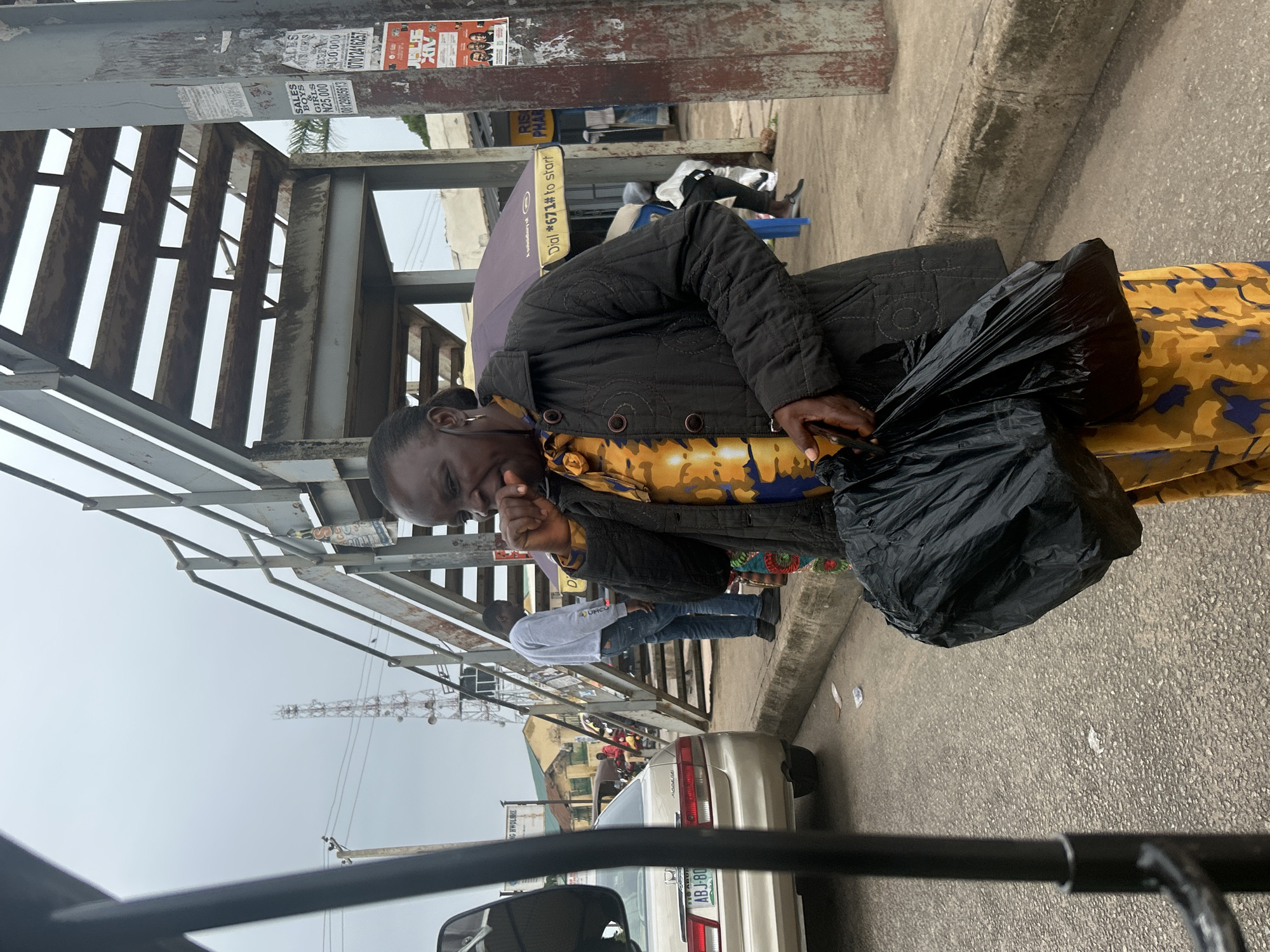 A medium shot depicts an elderly woman in Jos, Nigeria, standing on a roadside near a pedestrian bridge. She is wearing a patterned yellow dress, a dark quilted jacket, and carrying a large black plastic bag. Her hand is raised to her mouth as if coughing. A gray sedan is parked behind her, with a partial view of its license plate, which reads "ABJ-80 NG ABUJA". A man is walking on the pedestrian bridge. Utility poles and buildings are visible in the background. The setting appears to be a busy street in Jos.