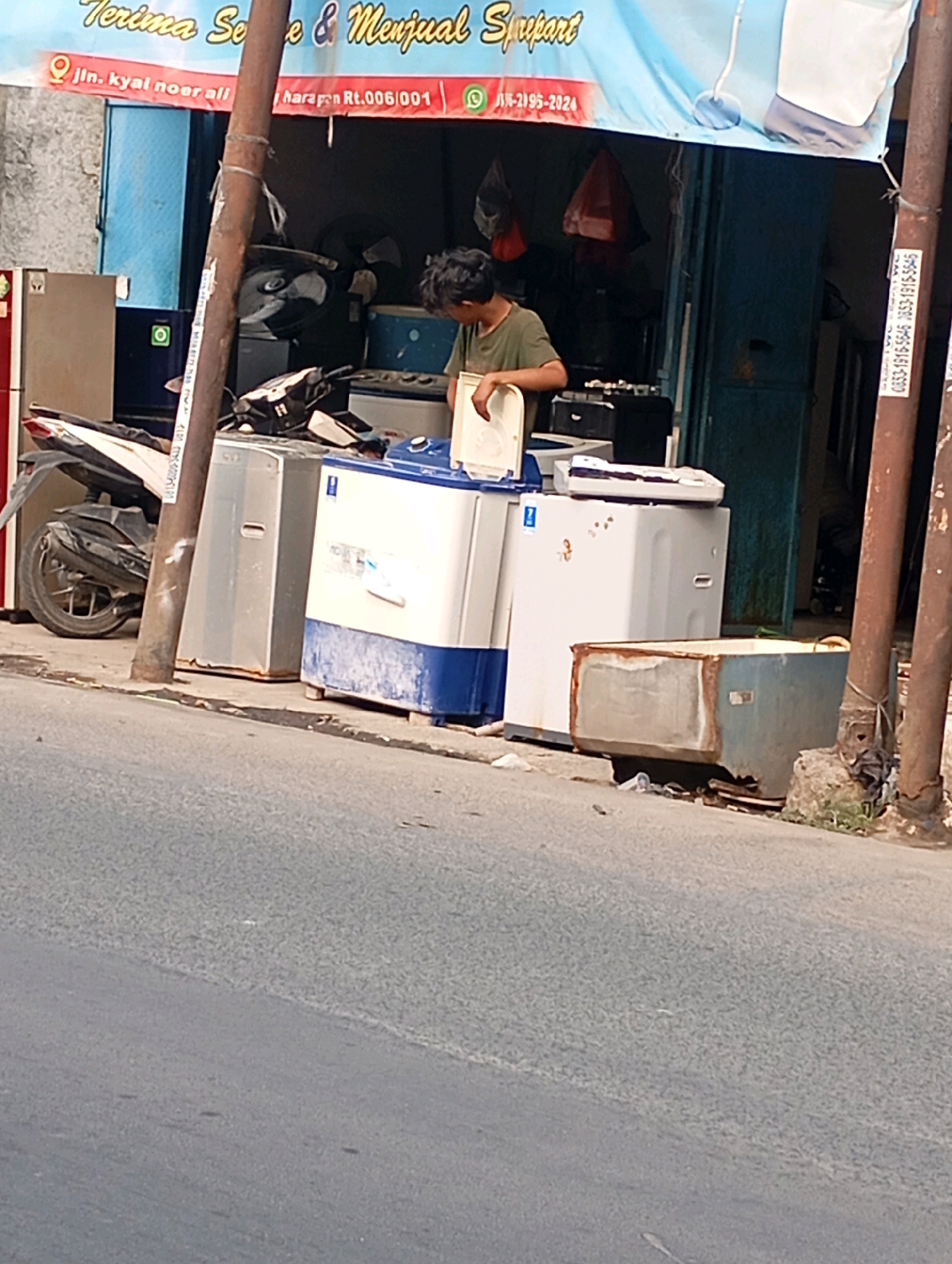 This image depicts a street scene in Babelan, Indonesia, likely near a store selling used appliances. In the foreground, a grey asphalt road takes up the lower half of the frame. Behind the road, on the sidewalk, several large appliances are visible, including washing machines and what appears to be a refrigerator. A person, a young male with dark hair, is standing amidst these appliances, appearing to be examining or working with one of the washing machines. A motorcycle is parked to the left of the appliances. Above the scene, a blue banner with text is stretched across the storefront, indicating the business name or services. The background shows the entrance to a building, possibly a shop, with various items visible inside. Two wooden poles, one thicker than the other and wrapped with what looks like tape or wire, stand vertically in the mid-ground. One of the poles has a vertical sticker with text. The overall environment suggests a commercial area with a focus on second-hand goods.