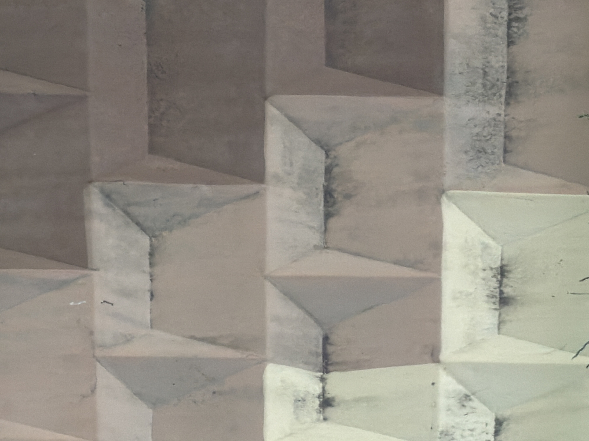 The image displays a close-up view of a weathered concrete barrier, characterized by its repeating geometric pattern of raised and recessed diamond-shaped sections. The concrete exhibits signs of age and exposure, with a mottled surface that includes areas of discoloration, staining, and some minor chipping. The color palette is muted, consisting of various shades of grey, beige, and a hint of light blue on some of the recessed surfaces. The lighting appears to be overcast, creating soft shadows that accentuate the three-dimensional texture of the barrier. The overall impression is one of utilitarian construction, typical of urban infrastructure. This type of barrier is commonly found in Minsk, Belarus, used for traffic control or to delineate spaces. There are no people or specific identifying landmarks visible in the frame.