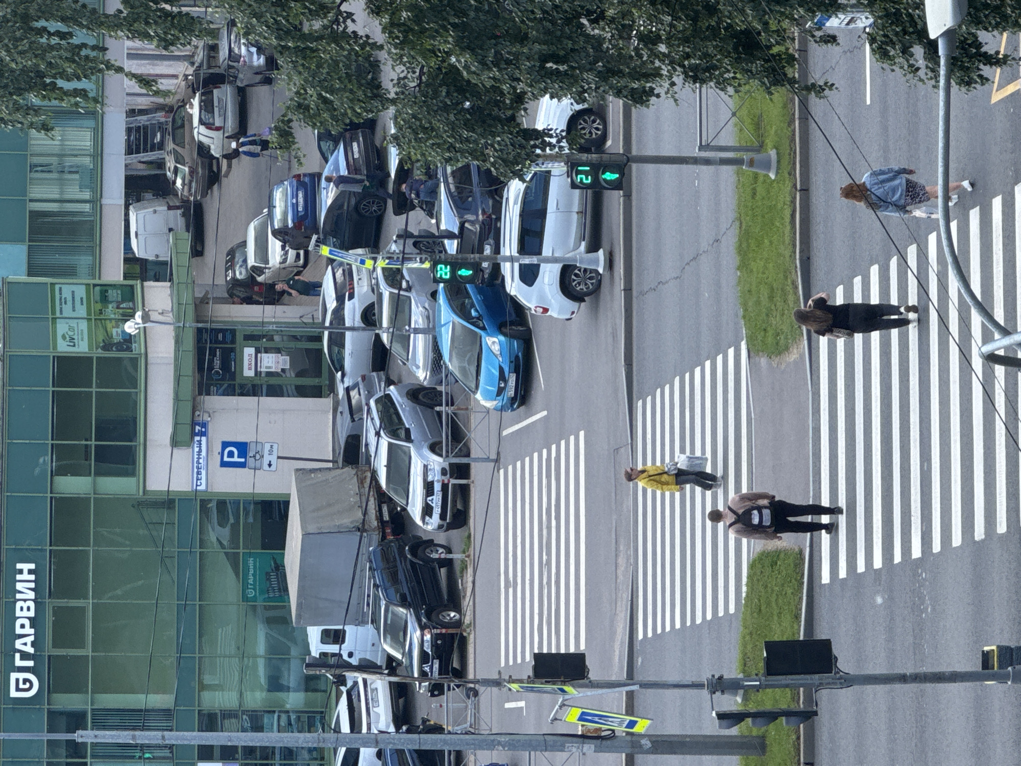 The image depicts a street scene in Saint Petersburg, Russia, with a focus on a pedestrian crossing. In the foreground, three pedestrians are visible crossing the street from right to left. The leftmost pedestrian is a woman with dark hair, wearing a black top and pants, with her back to the camera. To her right, another woman in a denim jacket and a patterned skirt is also crossing. A man with a backpack, wearing a light brown sweater and black pants, is further up the pedestrian crossing, also with his back to the camera.
Behind the pedestrians, a busy street with several cars is visible. The cars include a blue hatchback, a silver SUV, and a dark-colored SUV. A traffic light showing a green pedestrian signal is also present, with the number "21" displayed.
In the background, a multi-story building with green tinted windows dominates the scene. A sign on the building reads "GARVIN" in large white letters. Another sign indicates "SEVERNY 7" and a blue parking symbol with an arrow. Further into the building, the word "GARVIN" is also visible on a green sign above what appears to be a garage entrance. Several other vehicles, including vans and trucks, are parked or in motion within the building's vicinity. Trees with green foliage are present on the right side of the image, partially obscuring the view of the street. The overall impression is that of an urban environment during daylight hours.