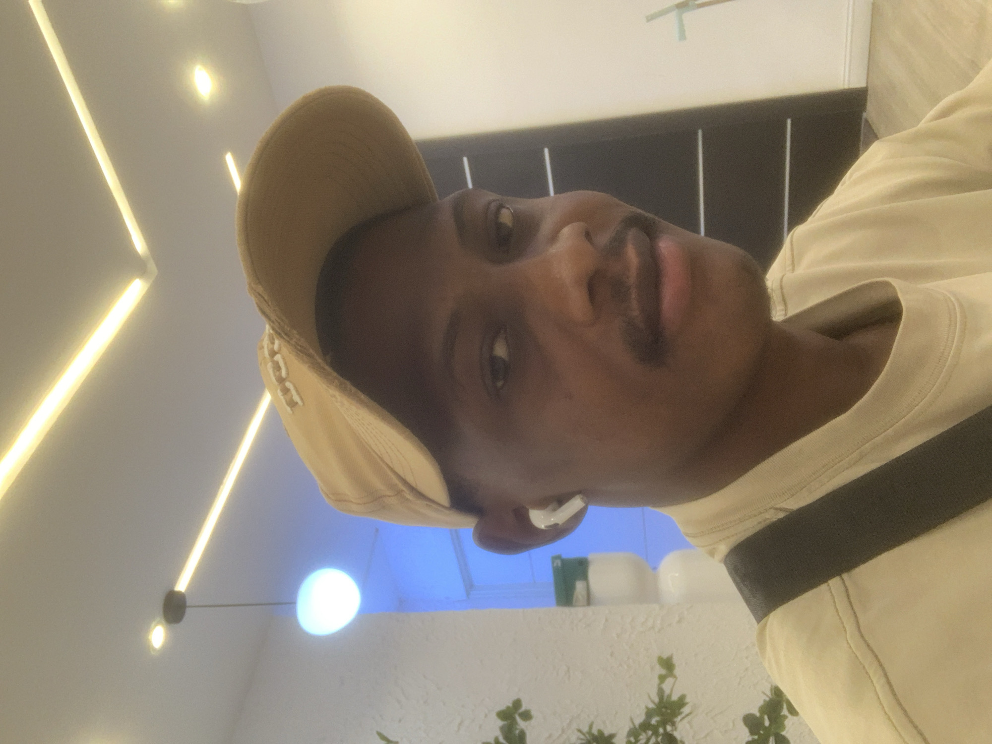 The image captures a close-up, angled portrait of a young man, presumably indoors. The man has a dark complexion and is looking directly at the camera with a neutral to slightly smiling expression. He is wearing a light beige or tan baseball cap, a light-colored (possibly cream or beige) t-shirt, and a dark shoulder bag strap diagonally across his chest. A white wireless earbud (AirPod) is visible in his right ear. He has a mustache and a faint goatee.
The background shows a modern interior setting. The ceiling features warm-toned linear LED strip lights and a suspended spherical light emitting a soft glow. Further in the background, a distinct blue ambient light emanates, possibly from an accent light or a light-colored object illuminated by blue light. Below the blue light, there's a white, textured surface, possibly a wall or a decorative planter, adorned with some green foliage. A dark, horizontal structure, perhaps a partition or a piece of furniture, is visible behind the man's head and shoulder. The overall lighting suggests an artificial indoor environment. There is no visible text in the image. The image is presented at an angle, making it appear as if the man's head is tilted to the right. The activity appears to be posing for a photograph. The location context is Animashawun, Nigeria.