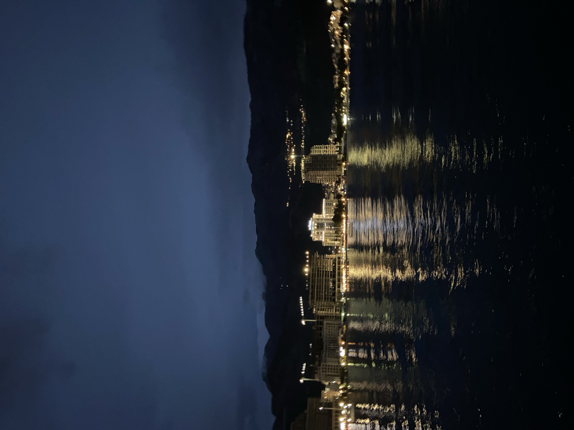 The image depicts a nighttime scene of Budva, Montenegro, as viewed across the water. The cityscape is illuminated with lights that reflect on the surface of the water. Buildings and hotels are visible along the coastline, stretching between the shore and a dark mountain range in the background. The sky is dark, suggesting either a clear night or heavy cloud cover. The overall impression is that of a quiet, nocturnal view of a coastal resort town.