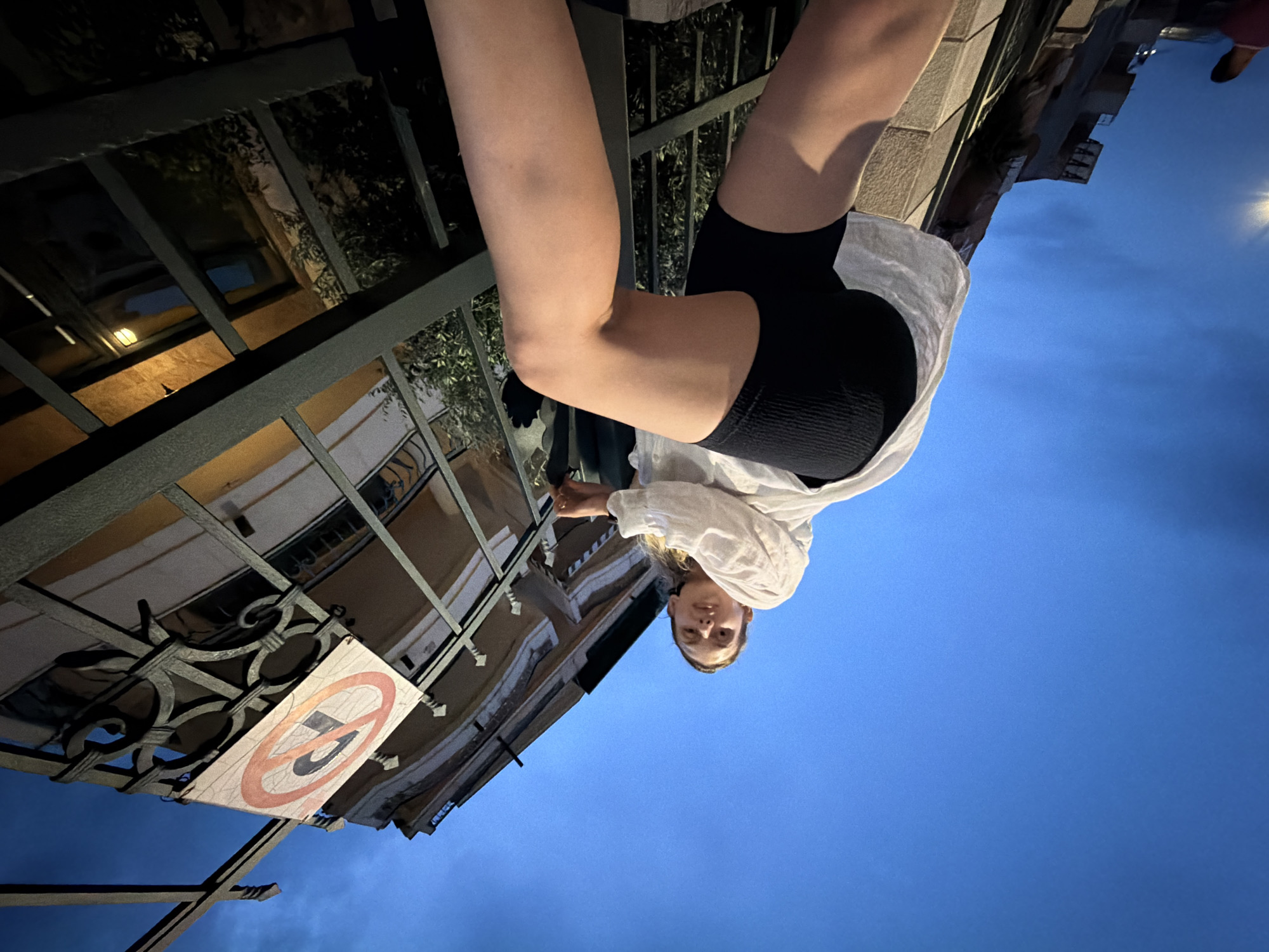 In Budva, Montenegro, a low-angle shot captures a young woman with blonde hair, wearing a white linen shirt and black biker shorts, posing against a backdrop of a dark blue evening sky. She is positioned on a ledge, with her legs extended and one arm bent, touching a decorative metal railing. A circular traffic sign with a red border and a red diagonal line through a black symbol, possibly indicating "no entry" or a similar prohibition, is attached to the railing above her. The scene includes architectural elements of a building with windows and an awning, as well as some foliage. The overall lighting suggests dusk or twilight.