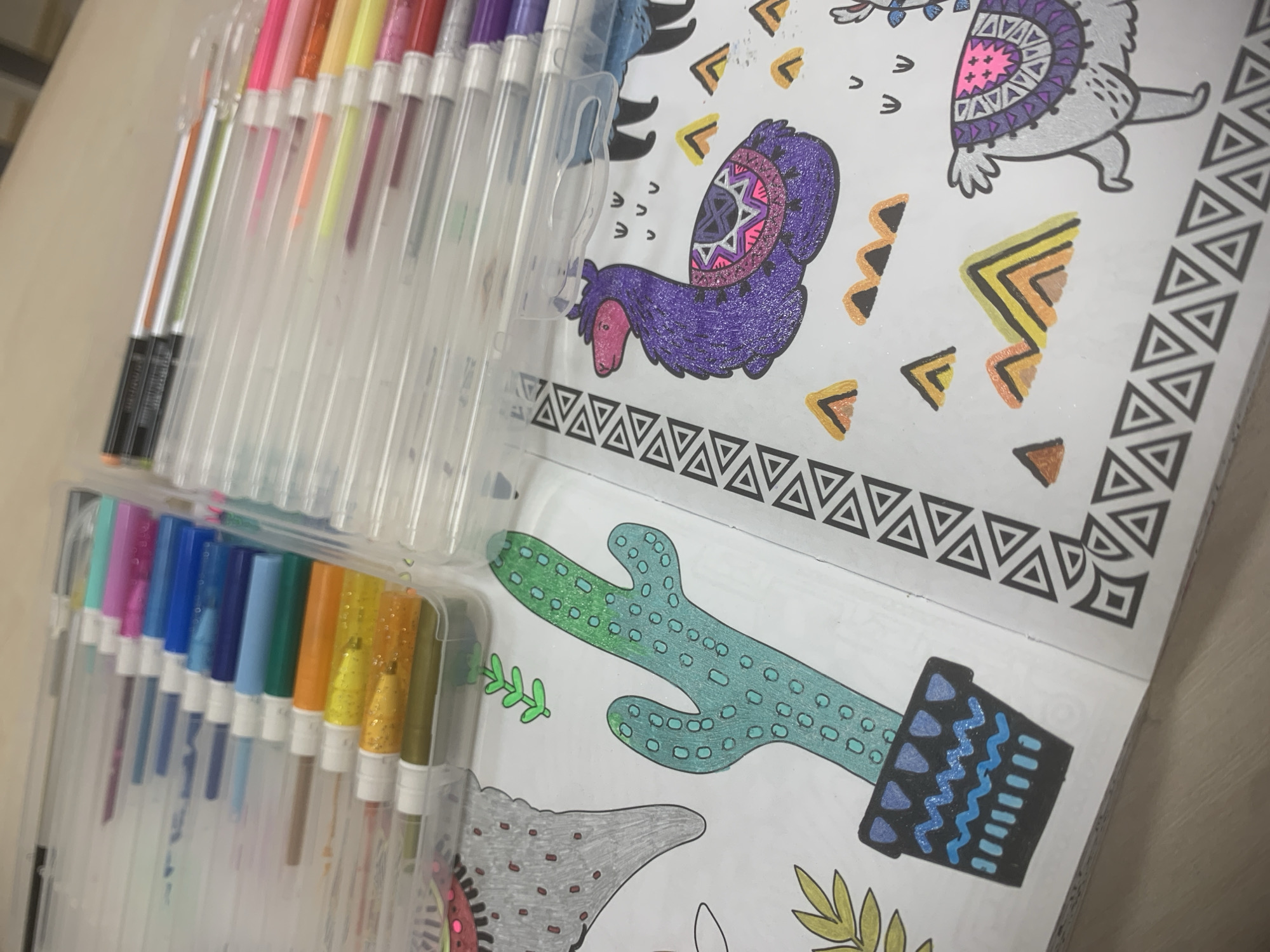 The image shows a coloring book with a set of colored pens. The coloring book features illustrations of llamas, a cactus in a pot, and decorative patterns. The illustrations are partially colored. The colored pens are in a clear plastic case, arranged by color. The background is a plain, light-colored surface. There is no indication of the image's location.