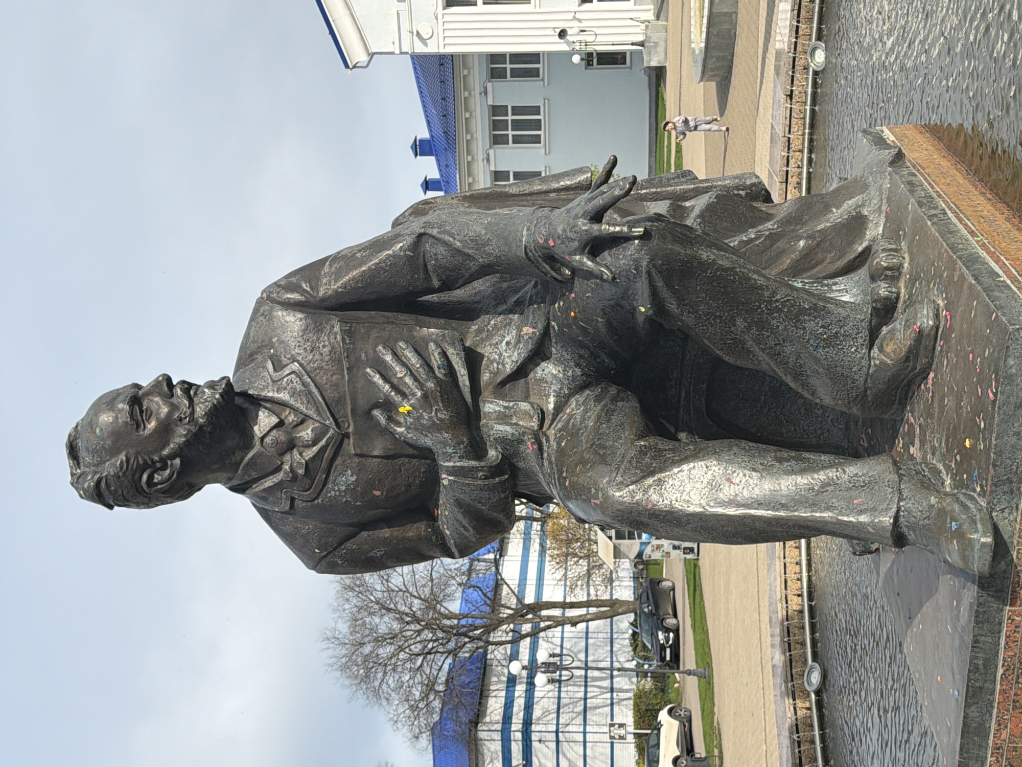 The image shows a bronze statue of a man seated on a platform, located in Chaikovsky, Russia. The statue is positioned in front of a building with white walls and a blue roof. There is a tree behind the building and a street with cars parked along the side. A woman is seen walking along the sidewalk in the background. The statue has a fountain running next to it.