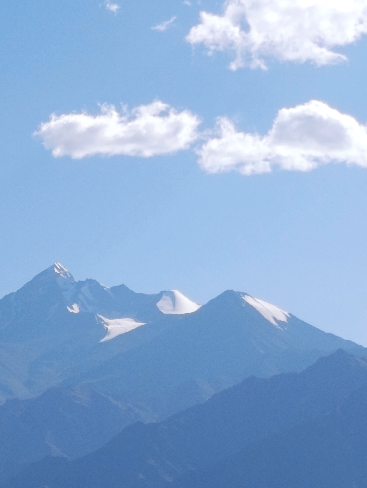 A serene landscape image depicts snow-capped mountain peaks under a clear blue sky dotted with scattered white clouds. The mountains are layered, with the closest peaks displaying rugged terrain and patches of snow, while the distant ones fade into a hazy blue silhouette. The scene evokes a sense of natural grandeur and tranquility. The location is likely in Shey, India, a region known for its stunning Himalayan vistas.