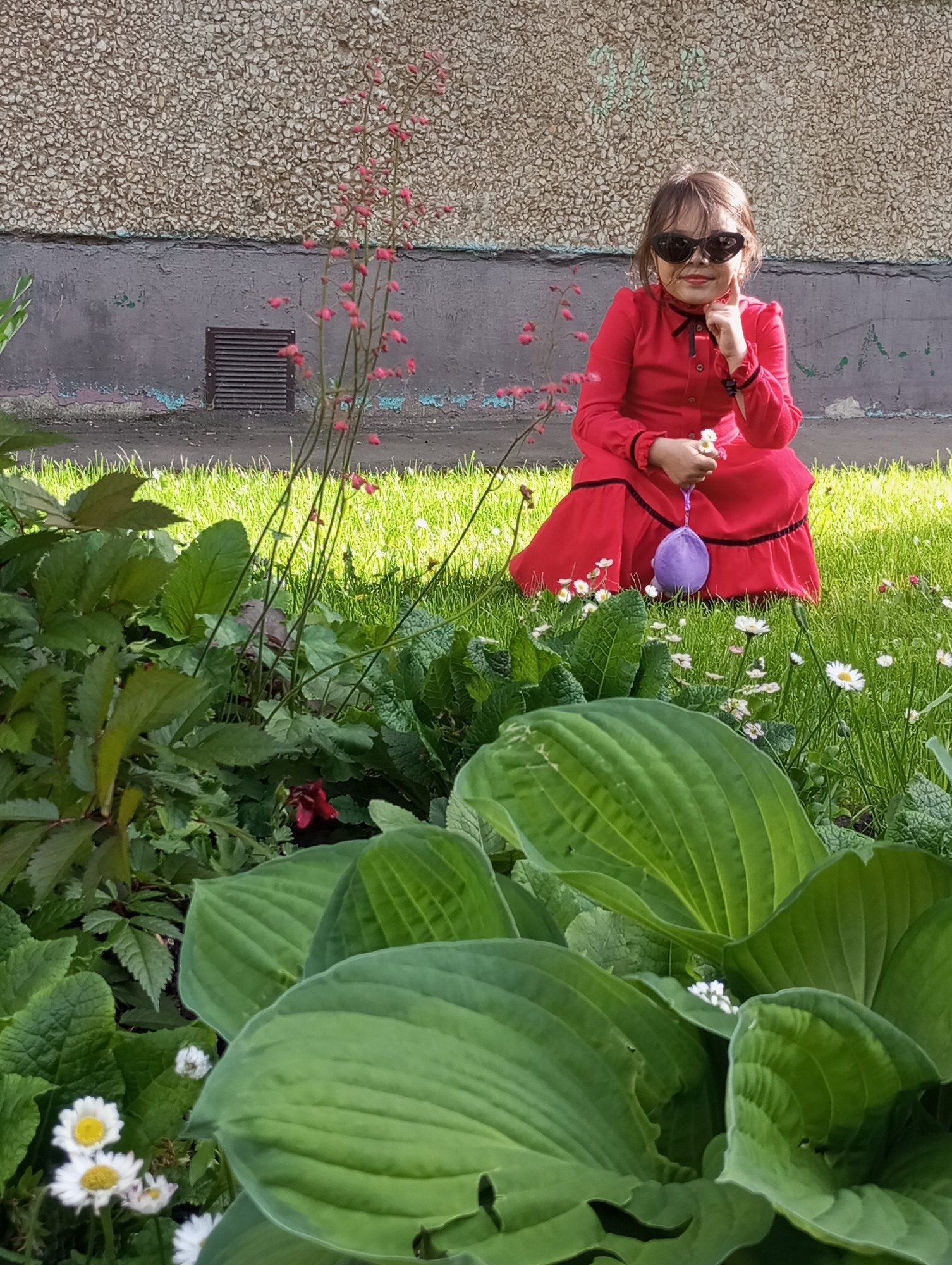 A young girl, likely a child, wearing a long-sleeved, collared red dress with black trim and buttons, kneels on a vibrant patch of green grass. She sports dark, heart-shaped sunglasses and looks directly at the camera, with her right index finger gently touching her cheek or chin. In her right hand, she holds a small white flower, and in her left, a purple, soft, balloon-like object.
The setting is an outdoor garden or green space, possibly in Izhevsk, Russia, adjacent to a building. The foreground is dominated by lush green foliage, including large-leafed plants (resembling Hostas) and clusters of small white daisy-like flowers, which also extend into the grass around the girl. A plant with slender stems and small pinkish-red flowers is visible to the girl's left.
In the background, a building wall rises. The upper section is textured, light brown or beige, suggesting a pebbledash or stucco finish, while the lower part is a smoother, greyish-purple concrete base showing signs of wear. A dark, rectangular ventilation grate is embedded in this lower wall section. The scene is brightly lit by natural sunlight, indicating a clear, sunny day, with strong shadows visible on the grass and plants. The girl appears calm and possibly playful, enjoying her time outdoors. There is no visible text in the image.