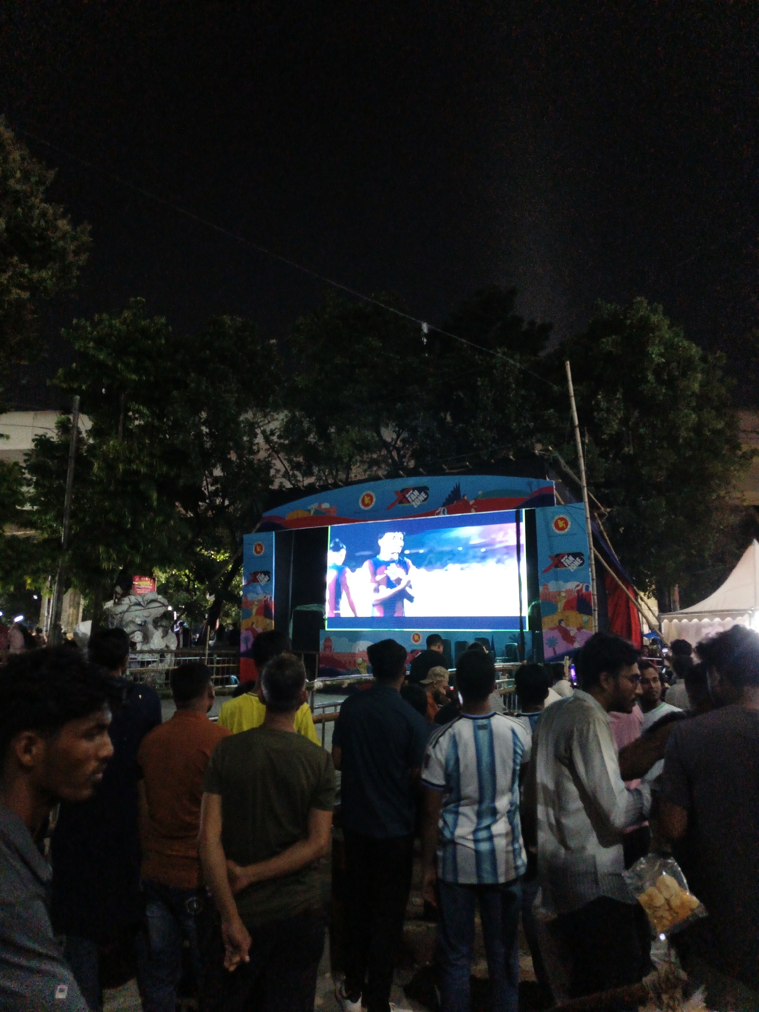 A crowd of people stands in the darkness of night, facing a large outdoor screen displaying a live broadcast of a sporting event. The screen is mounted on a brightly colored blue and red structure, likely a stage or booth, adorned with logos and cartoonish figures. Several individuals in the foreground are looking at the screen, some with their hands clasped behind their backs. The environment appears to be an open space, possibly a park or public square, with trees visible in the background and a white tent to the right. The overall scene suggests a community gathering to watch an event, with a festive yet casual atmosphere.