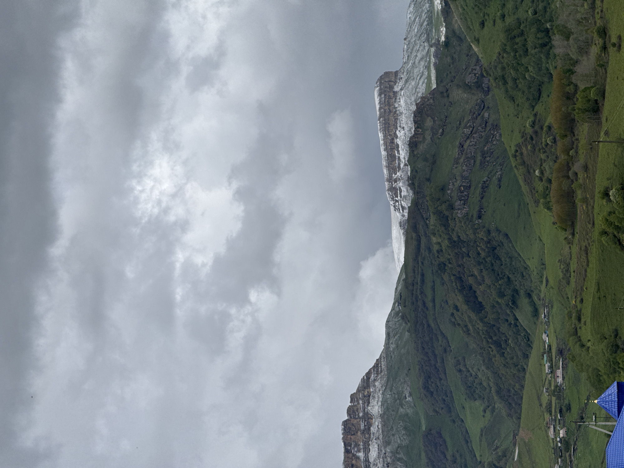 The photograph depicts a landscape scene in the city of Верхняя Мара, Russia. A tall, rocky mountain with patches of snow on its summit and along its ledges dominates the right side of the image. The mountain's slopes are covered in green vegetation. To the right of the mountain is a valley with a few scattered buildings and green pastures. The sky, which occupies the left side of the image, is overcast with grey clouds.
