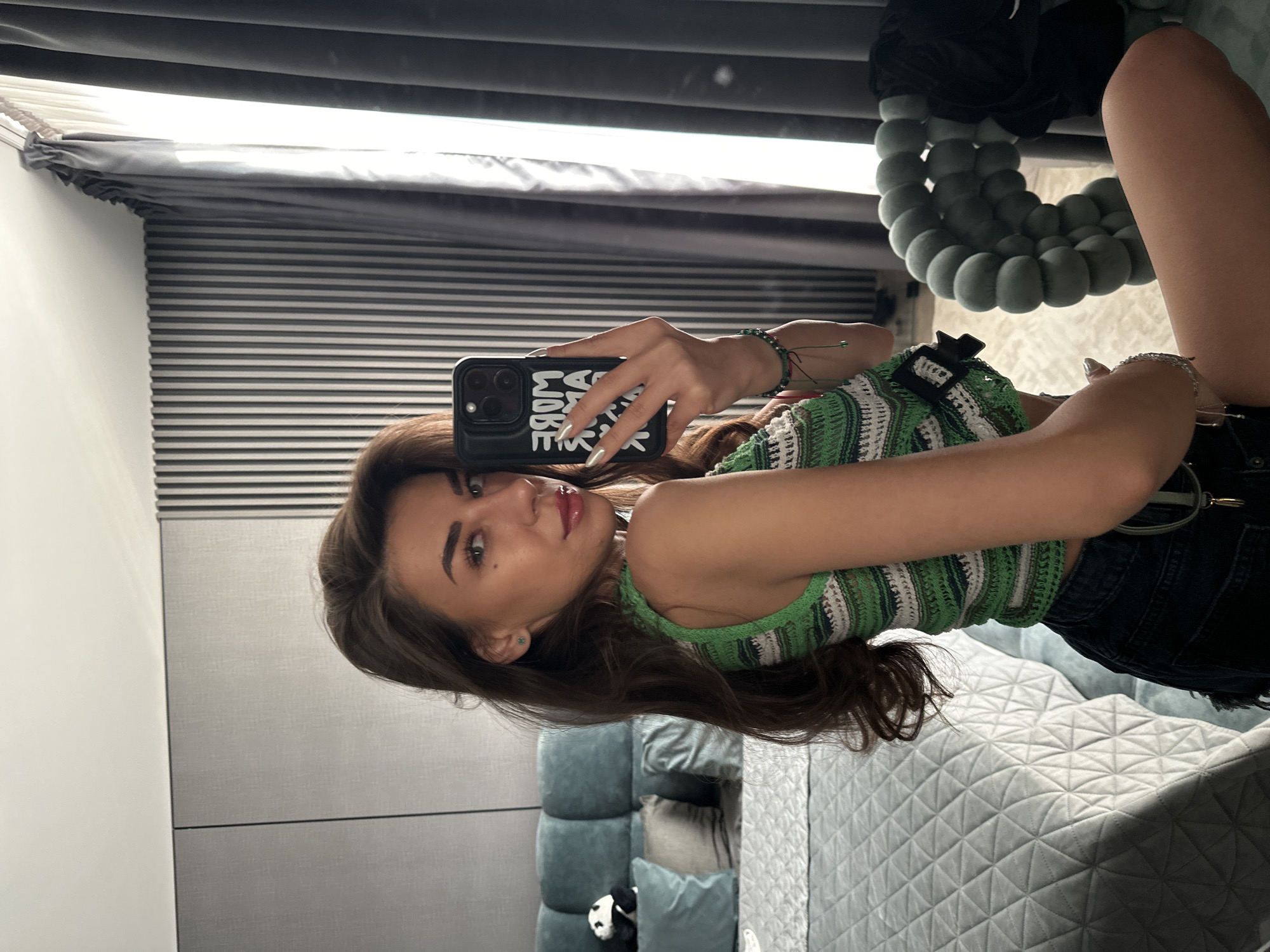 A young woman with long, dark brown hair is taking a mirror selfie in a bedroom. She is wearing a green and white striped crocheted tank top and dark denim shorts. Her phone, which has a black case with white lettering that reads "MORE AMBITIOUS," is visible in her right hand. She is wearing a small green earring and a beaded bracelet on her left wrist. In the background, a bed with a quilted grey comforter and several teal pillows is visible. A stuffed panda toy sits on the pillows. A textured grey wall with vertical slats is behind the bed, and dark grey curtains hang on the right side of the frame. A uniquely shaped, light grey chair with spherical cushions is in the foreground on the right. The setting appears to be a modern bedroom.