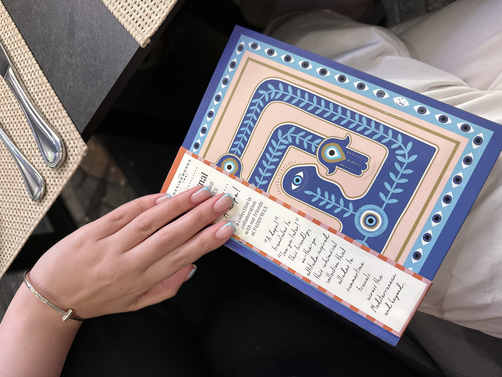 The image shows a person holding a Designworks Ink Paddywax book in their left hand. The book's cover features a pattern of eyes and hands in blue, pink, and gold. A white label is partially adhered to the book and shows some text.
The person is wearing a gold bracelet with a horizontal bar. They are sitting at a table, which is visible on the left side of the frame. The table has a gray top and a woven placemat. Cutlery, including a fork and a knife, are resting on the placemat. The person is wearing black pants and there is a white piece of fabric visible behind the book.
The image does not provide enough information to determine the specific location in Yerevan, Armenia.