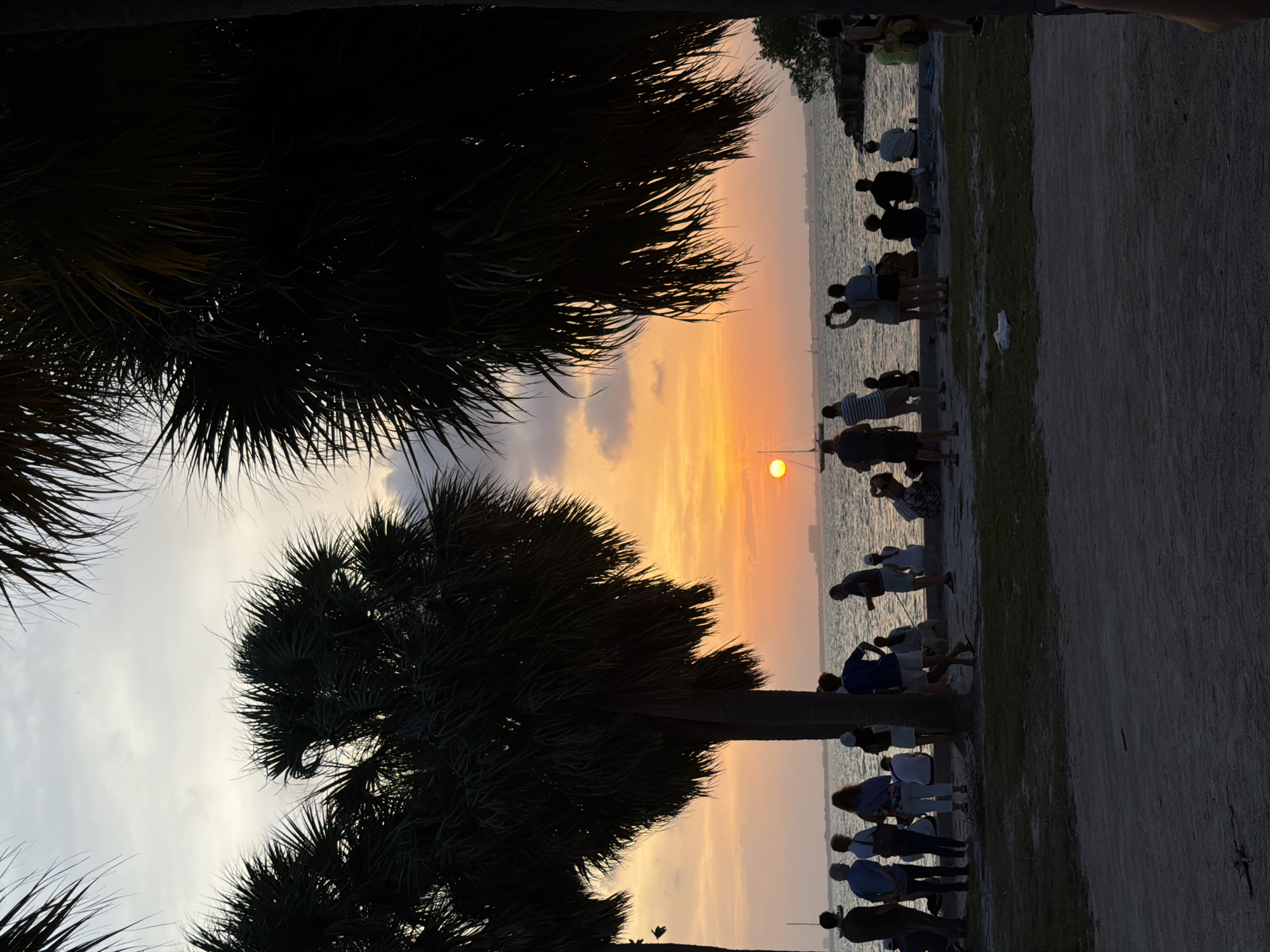 This image captures a sunset scene at the Bill Baggs Cape Florida State Park on Key Biscayne, United States. The sun, a bright orange orb, is low on the horizon, casting a warm glow across the sky and water. Silhouetted palm trees frame the scene, their fronds dark against the colorful sky. A group of people are gathered along the waterfront, some standing and others sitting, all facing the ocean to watch the sunset. A few sailboats are visible in the distance on the calm water. The foreground shows a sandy area and some grass, typical of a park setting. The overall atmosphere is peaceful and serene, with people enjoying the natural beauty of the coastal environment.