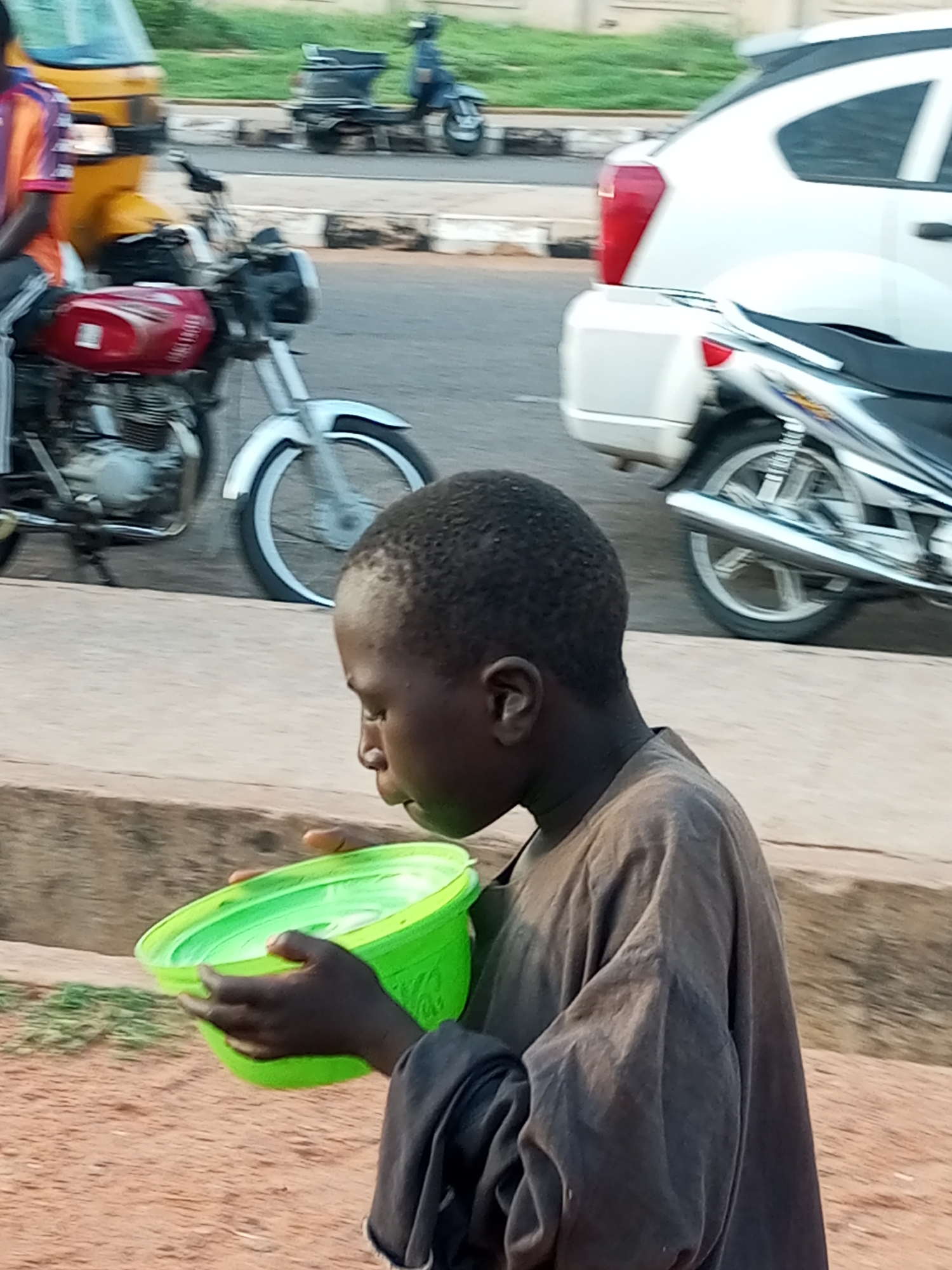The image depicts a street scene in Gombe, Nigeria, focusing on a young Black boy in the foreground. He is wearing a loose, dark brown garment and holding a bright green plastic bowl with both hands. His head is bowed, and he appears to be drinking or looking into the bowl. In the background, several vehicles are visible on a paved road. To the left, a yellow auto-rickshaw and a red motorcycle are parked. Further back, a white car and another motorcycle are also present. The environment suggests an urban setting with a sidewalk and grassy areas beyond the road. The lighting indicates it is daytime, likely late afternoon due to the shadows.