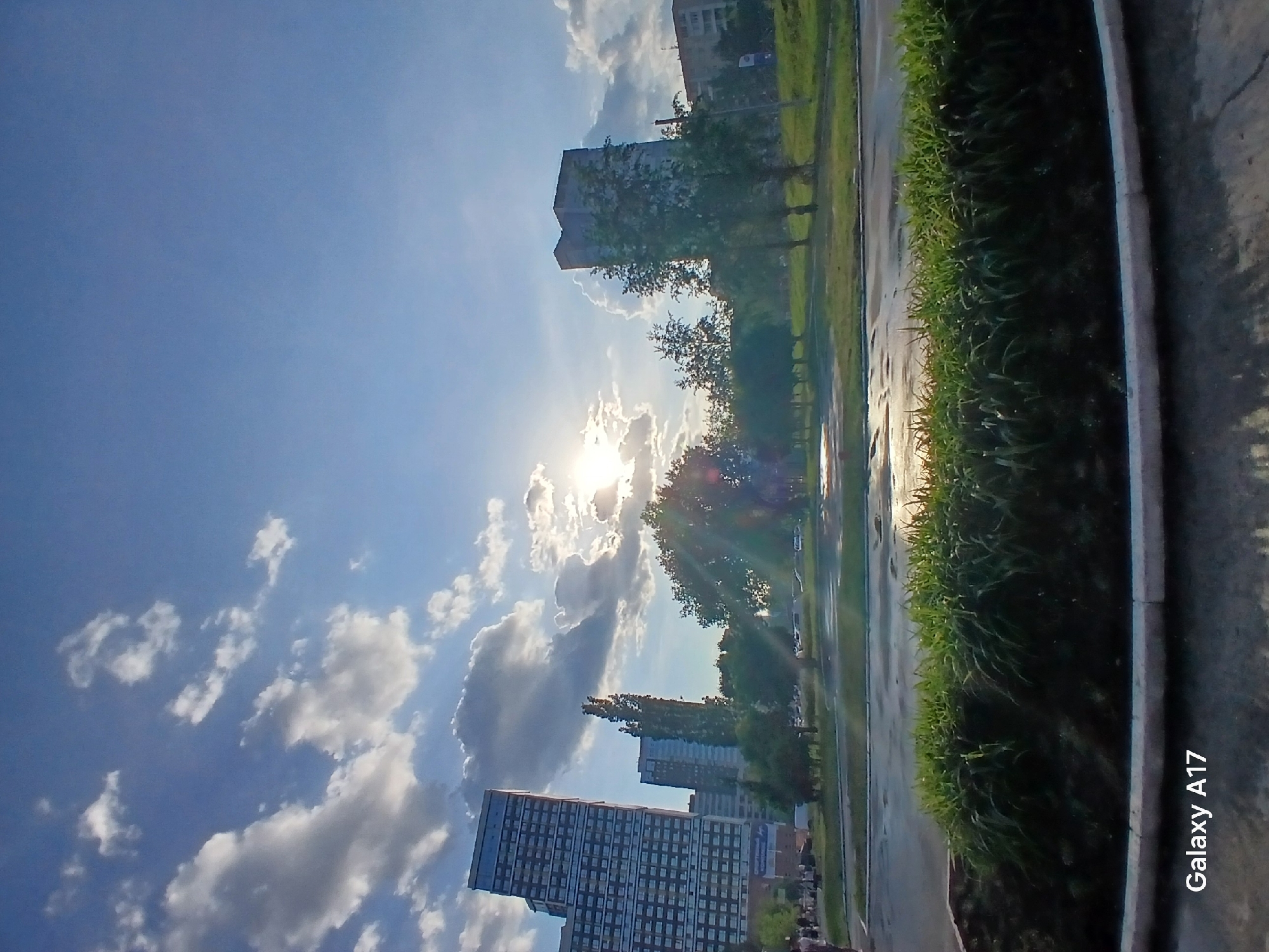 The image, taken in Yekaterinburg, Russia, shows a street scene with a view towards the sky. The sun is visible, partially obscured by clouds. Several tall buildings are present in the background. Along the right side of the image is a green strip of vegetation, possibly a line of bushes or a grassy area, next to a pavement. There are likely trees along the edge of the road. The sky is blue with scattered white clouds.