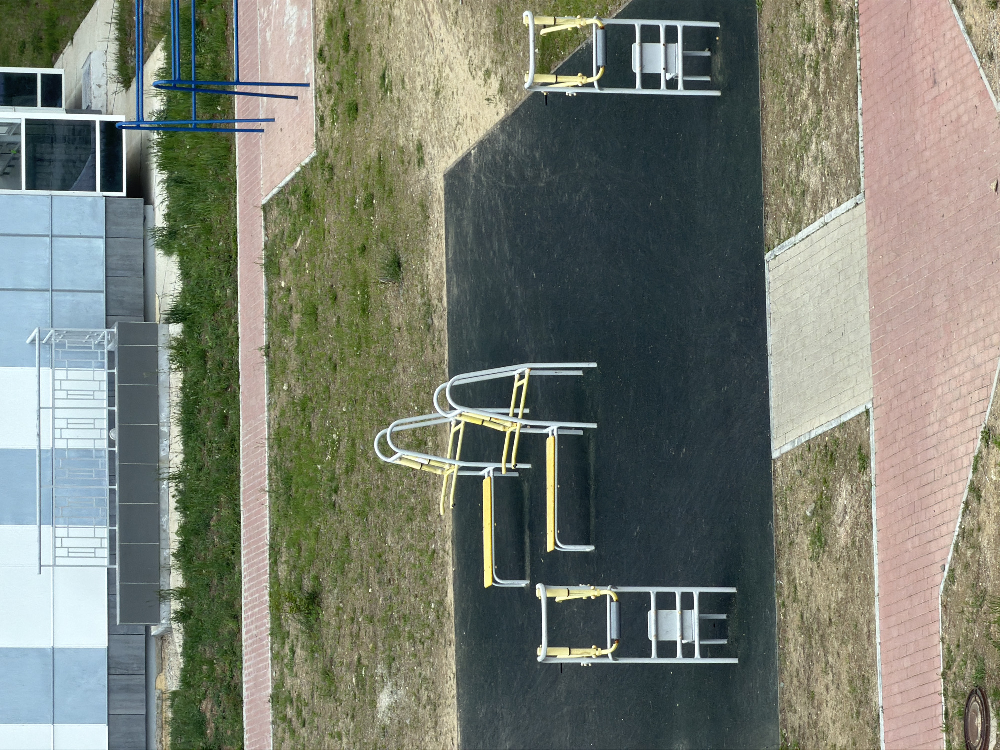 An overhead, high-angle view captures an outdoor exercise area. The scene is likely in Tsiolkovsky, Russia, given the context. The primary subject is a black rubberized surface, presumably for safety, on which several pieces of exercise equipment are situated. Two sets of exercise equipment, each featuring a yellow bench supported by a white metal frame, are positioned in the center of the black surface. To the left and right edges of the black surface are individual pieces of exercise equipment, also with white metal frames and yellow elements. The exercise area is bordered by a beige paved pathway and a red brick pathway, with patches of dry grass and dirt surrounding the paved areas. In the background, a modern building with a façade of alternating light blue and white squares and a metal railing is visible. To the far right, a blue metal structure, possibly part of another playground or recreational setup, is partially visible. No people are present in the image. The overall impression is that of an unused public recreational space.