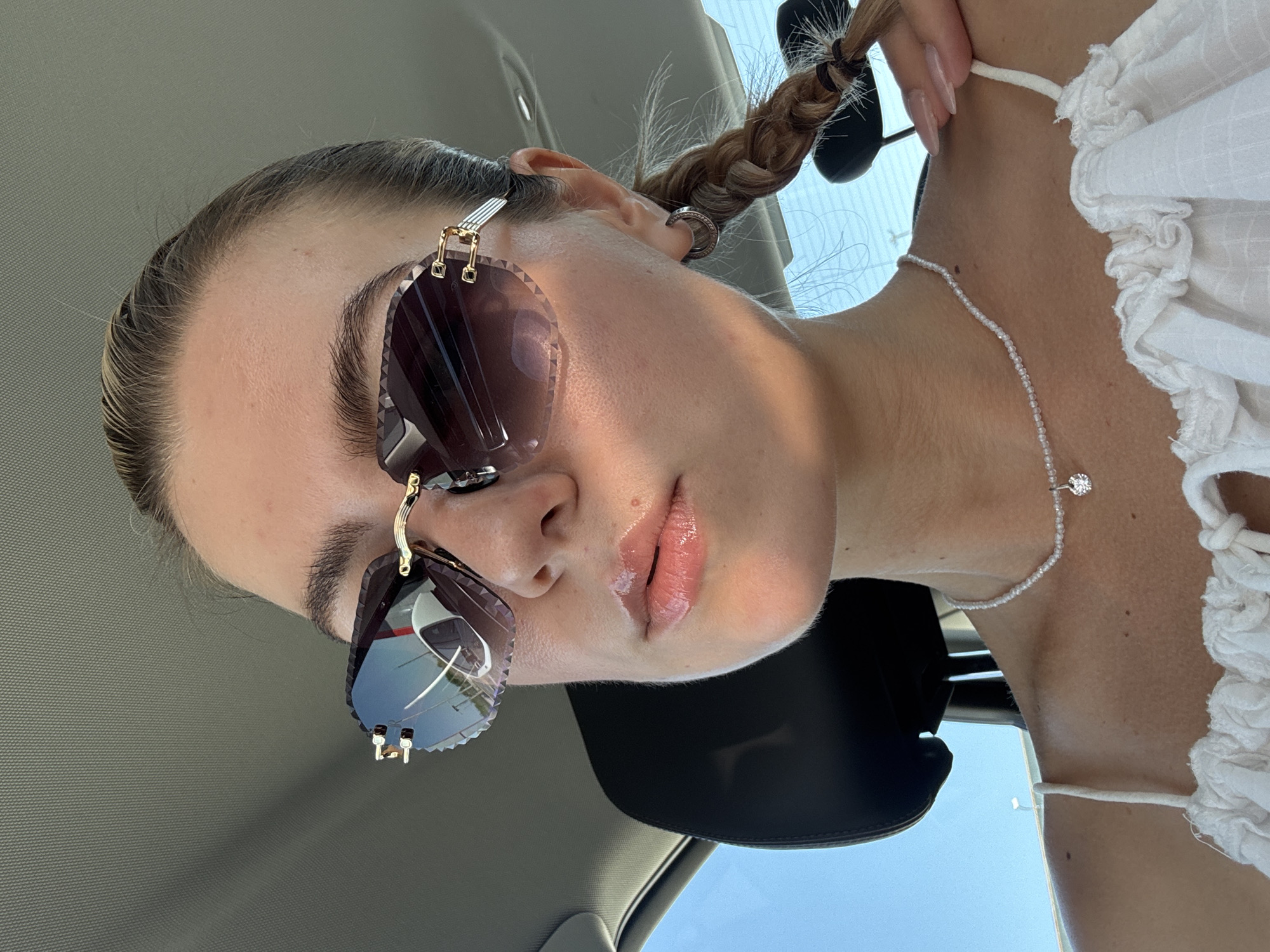 A female individual is positioned in a car, wearing sunglasses with a distinct rimless design. Her hair is styled in a braid. She is wearing a white top and a beaded necklace. The environment suggests a sunny day, as indicated by the sky visible through the car window.