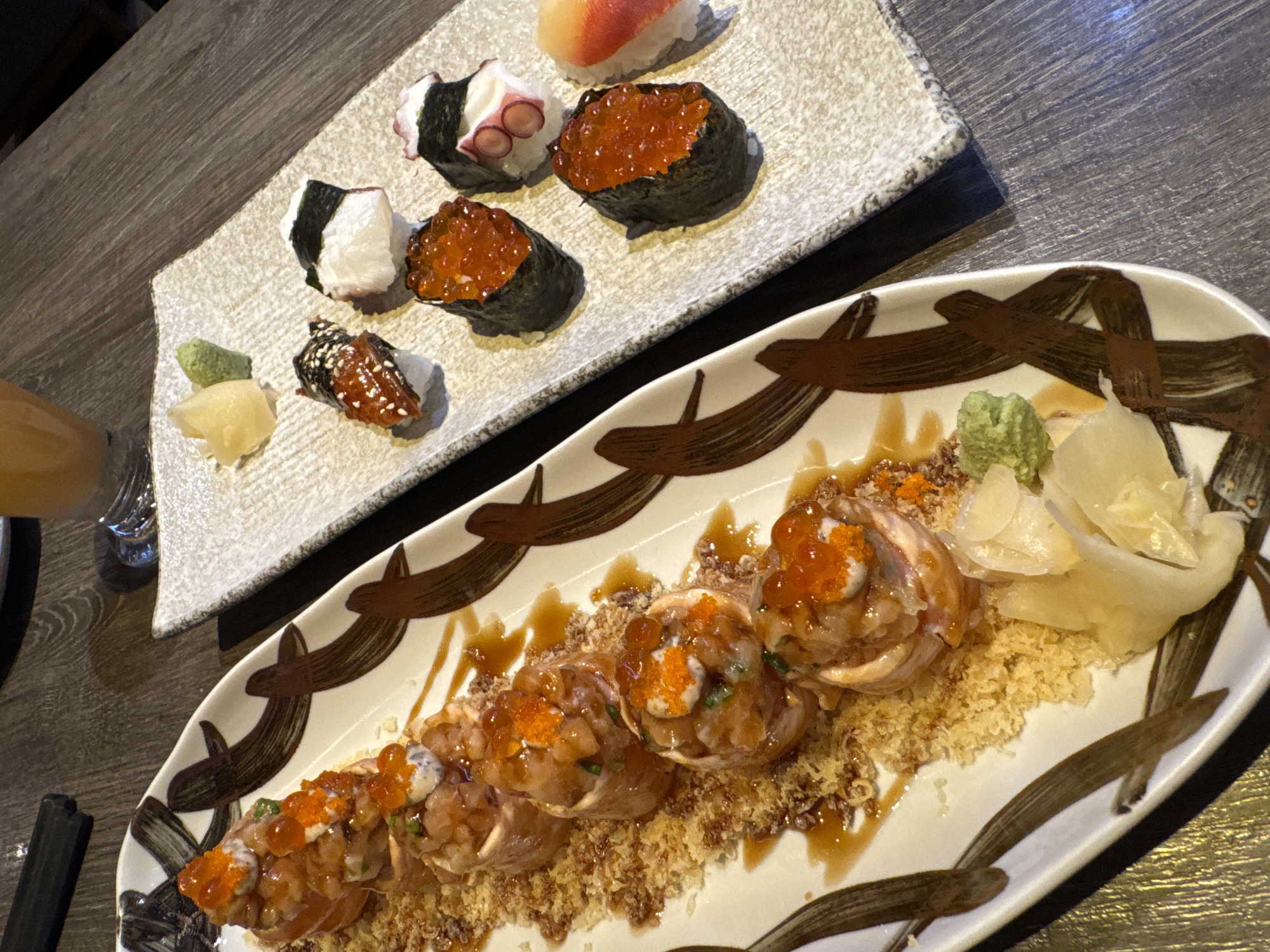 Two plates of sushi are on a wooden table. The top plate is rectangular and contains a variety of sushi, including a piece with red fish on top, one with octopus, several wrapped in seaweed and topped with orange roe, and a piece of eel. There's also a small dollop of wasabi and some ginger. The bottom plate is long and oval-shaped, with a decorative brown pattern. It features a row of sushi rolls covered in roe and a crispy topping, with wasabi and ginger on the side. A glass containing a yellow liquid is partially visible in the background on the left. A pair of chopsticks is also visible. The image suggests a meal at a restaurant in บ้านบางรักษ์, Thailand, although this can't be confirmed by the image alone.