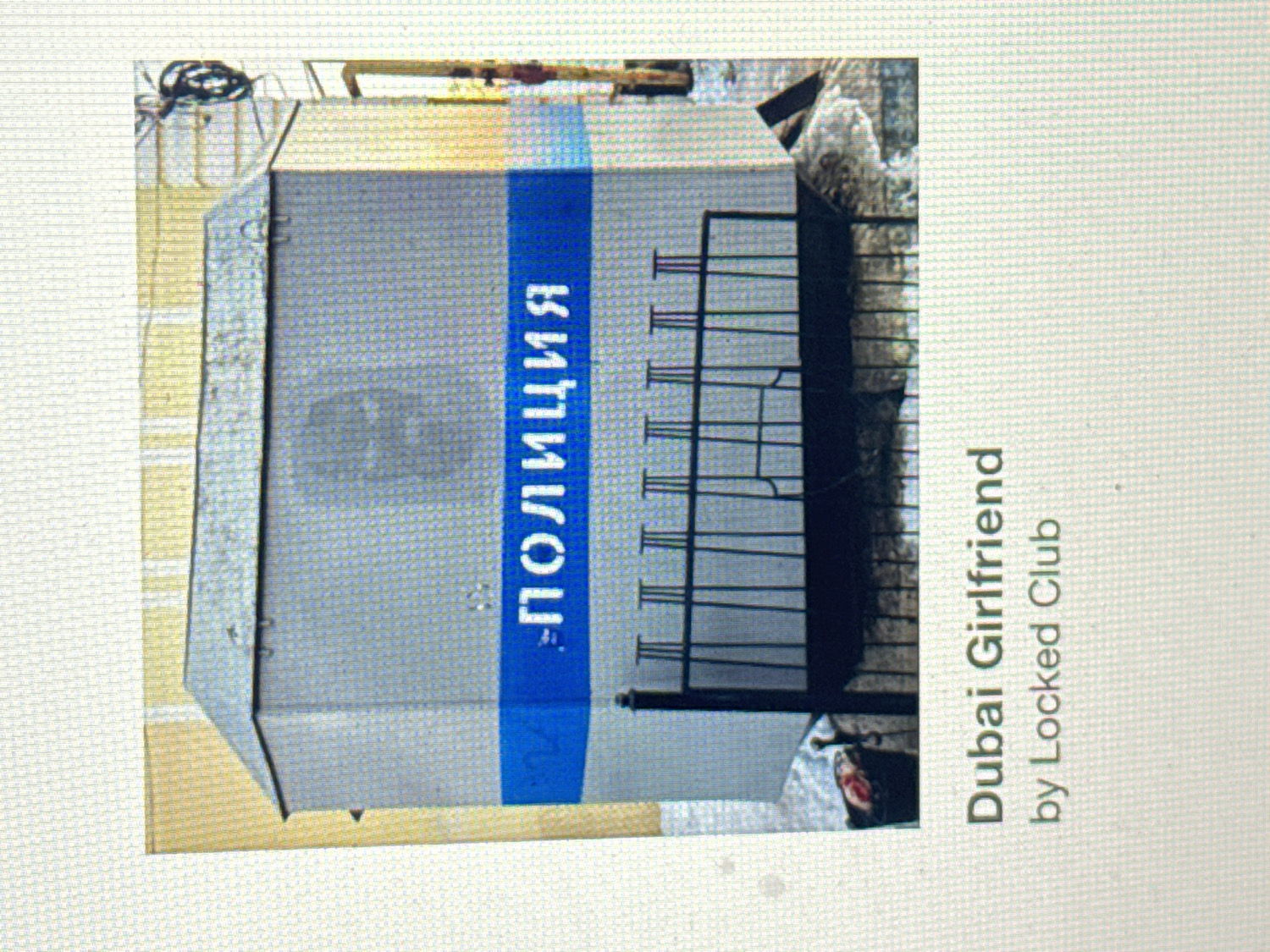 The image features a grey, rectangular, box-like structure, likely a kiosk, booth, or utility box, positioned outdoors. A vertical blue stripe runs down its front, bearing white Cyrillic text that clearly reads "ПОЛИЦИЯ" (POLITSIYA), which translates to "POLICE". The structure appears to be constructed from metal, showing visible seams and rivets.
To the right of this police box, a black metal rack is visible, consisting of several vertical posts and horizontal bars, possibly functioning as a barrier, a bicycle rack, or a support structure. The ground in the background appears somewhat uneven and cluttered, hinting at an outdoor setting that could be urban or industrial, with some debris or rough terrain visible. The lighting suggests it is daytime.
No people are visible in the scene, and no specific activity is taking place; the objects are stationary.
Overlaid on the lower right side of the image, rotated vertically, is additional text in white against a grey background. It reads: "Dubai Girlfriend by Locked Club".
Given the Cyrillic text "ПОЛИЦИЯ" and the provided location context, the scene is situated in Krasnoyarsk, Russia.