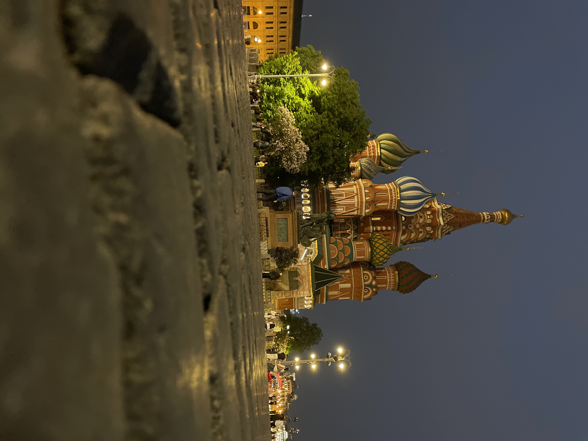 The image shows Saint Basil's Cathedral in Moscow, Russia, at night. The view is from ground level, with cobblestones in the foreground. The cathedral's colorful onion domes and spires are prominently displayed. A monument and trees are located near the cathedral. People are visible near the monument and along the street. Streetlights illuminate the scene. The sky is a dark blue, suggesting dusk or night. Buildings are visible in the background.
