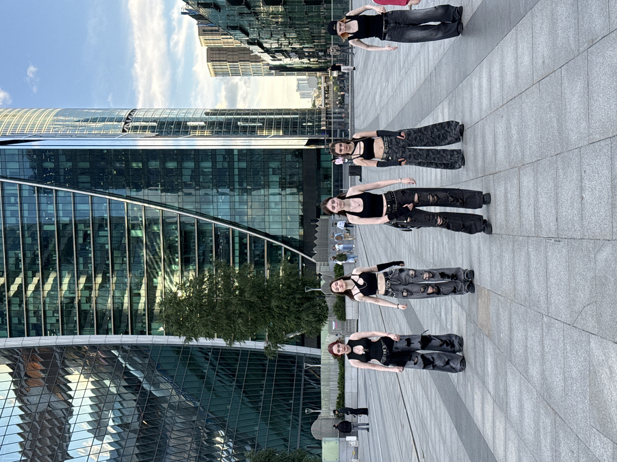 Five young women stand in an urban plaza in front of modern skyscrapers. The women are dressed in similar dark, baggy, ripped pants and black crop tops. The plaza has a patterned stone pavement, and in the background, there are large, reflective glass buildings, trees, and a cloudy blue sky. Some pedestrians are visible in the distance.