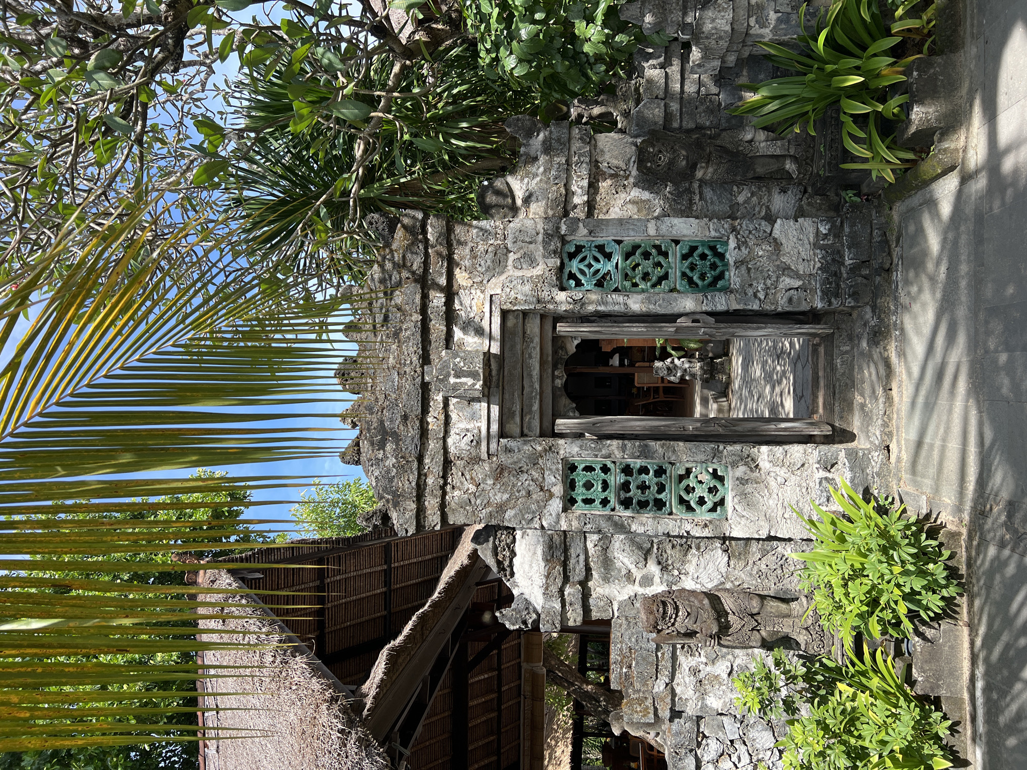 This image depicts a traditional stone building, likely a temple or part of an ancient complex, nestled within a lush tropical environment in Denpasar, Indonesia.
The central subject is a rustic building constructed from rough-hewn grey stones, exhibiting an aged appearance. A prominent arched doorway is centrally located on the facade, offering a glimpse into a dimly lit interior where some furniture can be vaguely discerned. Above and flanking the doorway are intricate, green-colored grilles or decorative vents, which add a vibrant, ornate detail to the stone. To the left, a portion of a traditional thatched roof, likely made from palm fronds or straw, is visible, suggesting an adjoining structure.
The scene is heavily framed by abundant tropical vegetation. A large palm frond extends into the foreground from the left, partially obscuring the view. Various leafy green trees and shrubs, including what appear to be frangipani trees with their characteristic branches, grow around and atop the stone walls, integrating the architecture with nature. To the right of the doorway, a partially visible, intricately carved stone statue, possibly a guardian figure or deity, stands, showcasing typical Indonesian artistry. A paved pathway or floor extends from the right side of the building.
The bright, clear blue sky and pronounced shadows indicate a sunny daytime setting, likely late morning or early afternoon. No specific activity is taking place; the scene is static, highlighting the architectural and natural beauty. No visible text is present. The overall atmosphere is one of serene history and natural charm, characteristic of Balinese landscapes and structures.