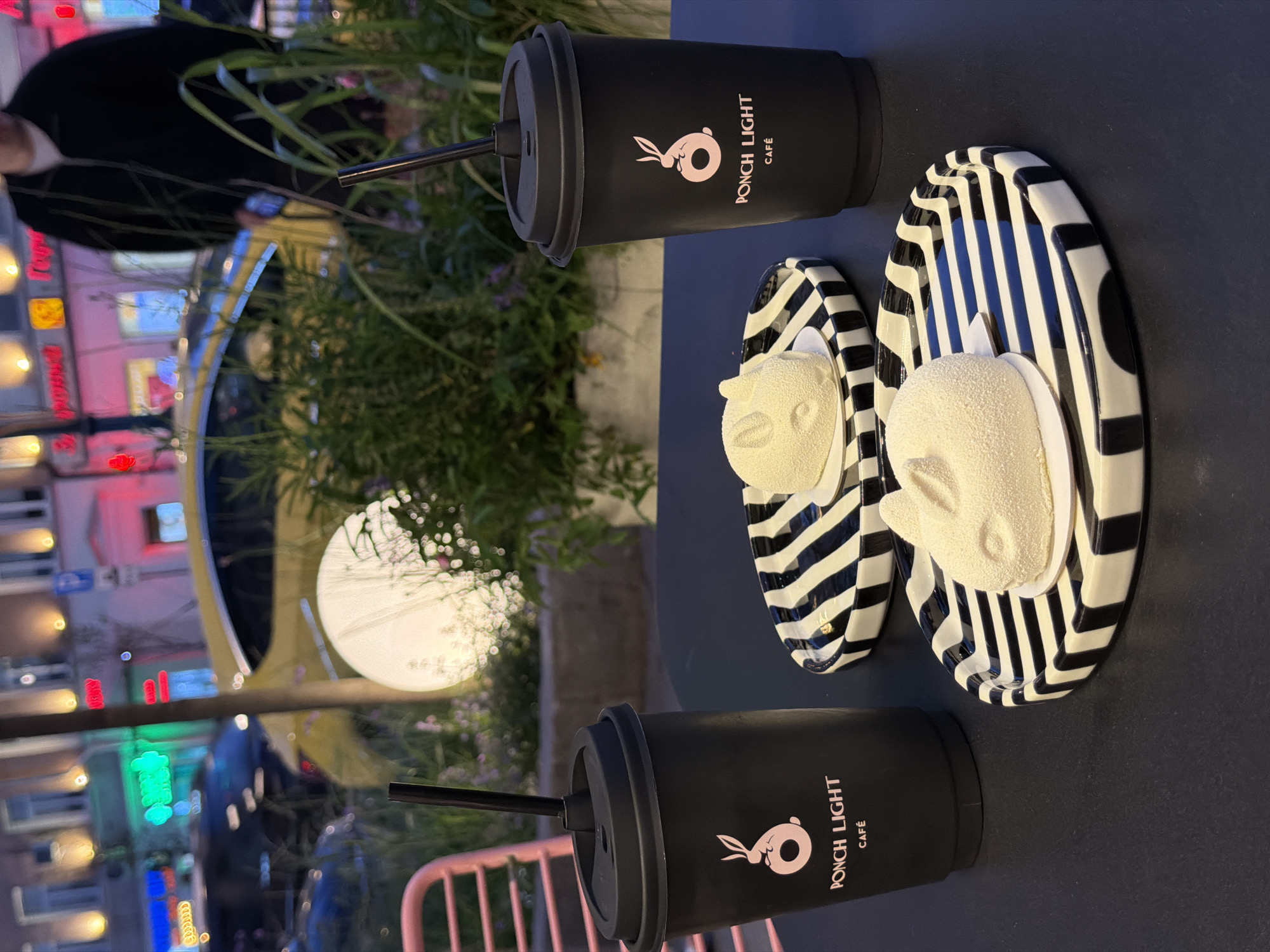 The image displays two black coffee cups with the text "PONCH LIGHT CAFE" and a pink bunny logo. Each cup has a straw. Next to the cups are two white desserts on black and white striped plates, shaped like animal faces. The desserts appear to be a light color and have a powdery texture. The scene is set on a dark surface, possibly a table, outdoors. In the background, there is greenery and a blurred reflection of buildings with colorful lights, suggesting an urban setting at night. A yellow car is partially visible in the reflection. A pink metal chair is also visible in the foreground. The overall mood seems relaxed and casual, possibly at a cafe or outdoor seating area. The time of day is likely evening or night due to the artificial lighting and reflections. The location context provided is Novaya Usman', Russia.