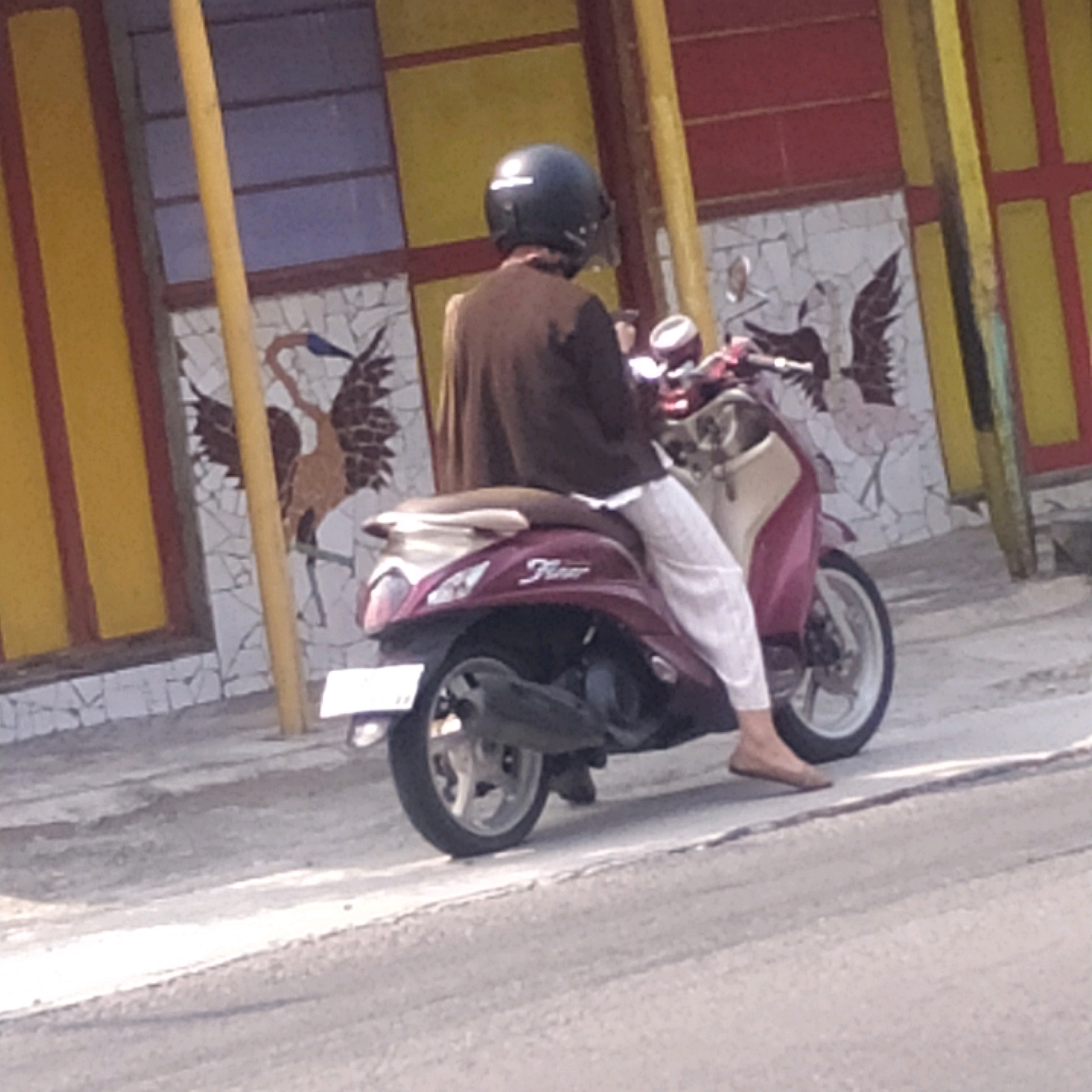 A person wearing a black helmet and a brown jacket is riding a dark red scooter. The scooter has "Fino" written on its side. The person is wearing white pants and brown flat shoes. The background shows a mosaic wall with bird designs and yellow and red pillars. The scene appears to be outdoors, possibly on a street or pathway in Batan, Indonesia.
