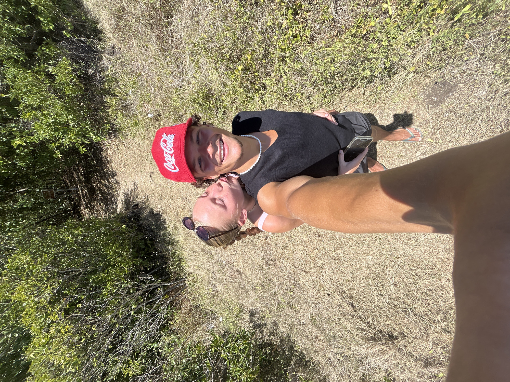 An elevated selfie captures two young adults interacting on a dry, unpaved path within a natural, vegetated environment, located in Le Gosier, France.
The primary subject, a male, is positioned centrally, facing the camera with a wide smile. He has curly brown hair and wears a red baseball cap with "Coca-Cola" in white script. His attire consists of a black, V-neck, sleeveless top and dark shorts. A white pearl-like necklace adorns his neck, and he wears light-colored flip-flops. A dark smartphone is held in his left hand, tucked against his side.
Behind and to the left of the male, a female subject leans in, kissing his right cheek with her eyes closed. She has blonde hair styled in a braid and wears sunglasses pushed up onto her head. A light-colored top is partially visible. Her left arm is wrapped around the male's waist.
The image is captured by an unseen individual, whose tanned forearm is prominently visible in the lower left foreground, holding the camera high.
The immediate ground consists of a path covered in dry, light brown grass and sparse green vegetation. The background features dense green bushes and trees, indicative of an outdoor, uncultivated area or trail. Bright sunlight casts distinct shadows, suggesting midday conditions. A small, indistinct wooden sign is partially visible within the foliage in the upper right background.