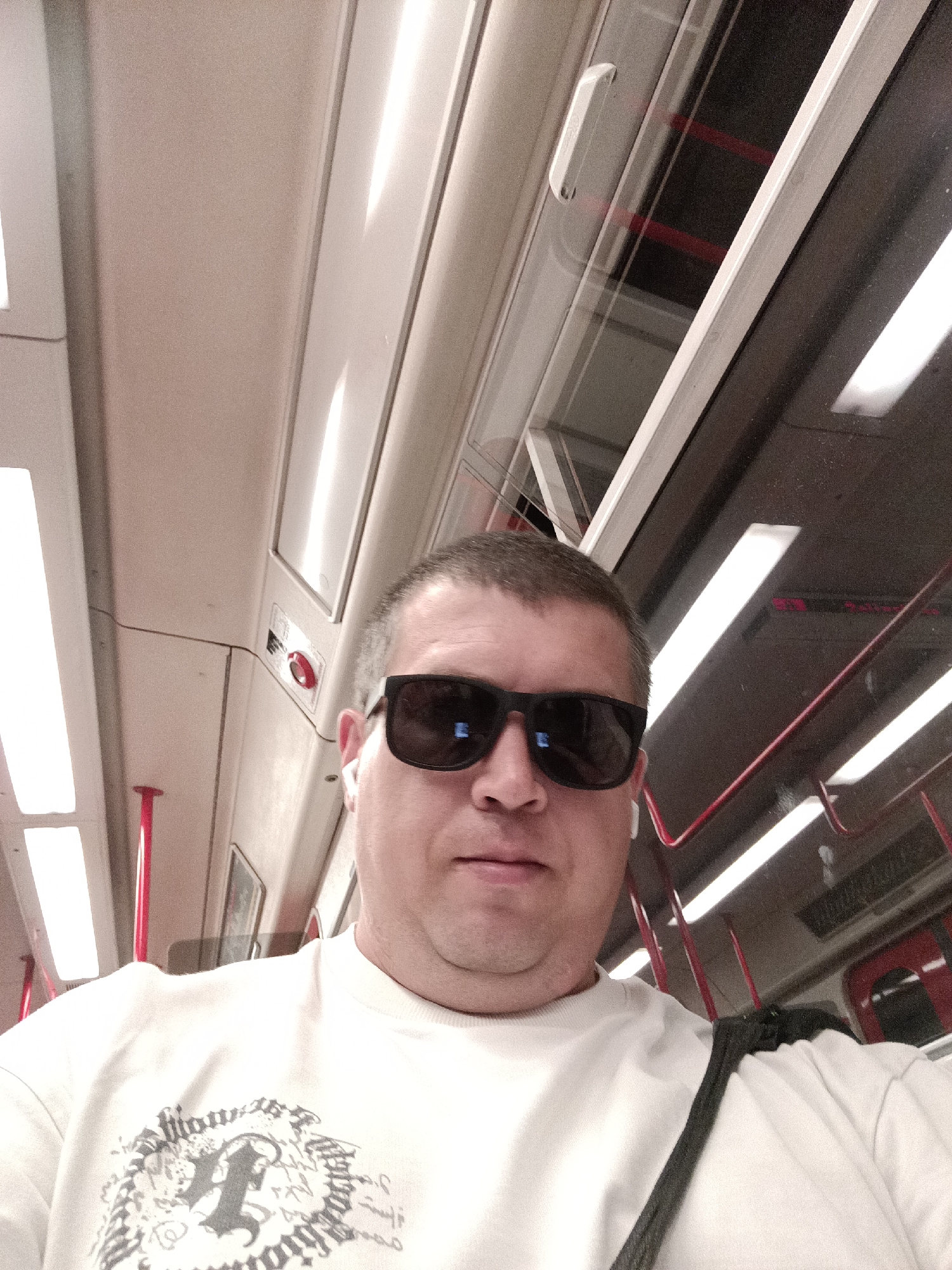 The media file is a selfie taken by a man wearing sunglasses and white earbuds, inside a Prague metro train. He is wearing a white sweatshirt with a black graphic print. The interior of the train car is visible, with white walls, ceiling lights, and red poles. The windows show a dark, reflective surface, possibly indicating the train is moving through a tunnel. The time of day is not discernible, and the weather is irrelevant as the image is taken indoors. The man's expression is neutral, and he appears to be relaxed. There is no visible text in the image.